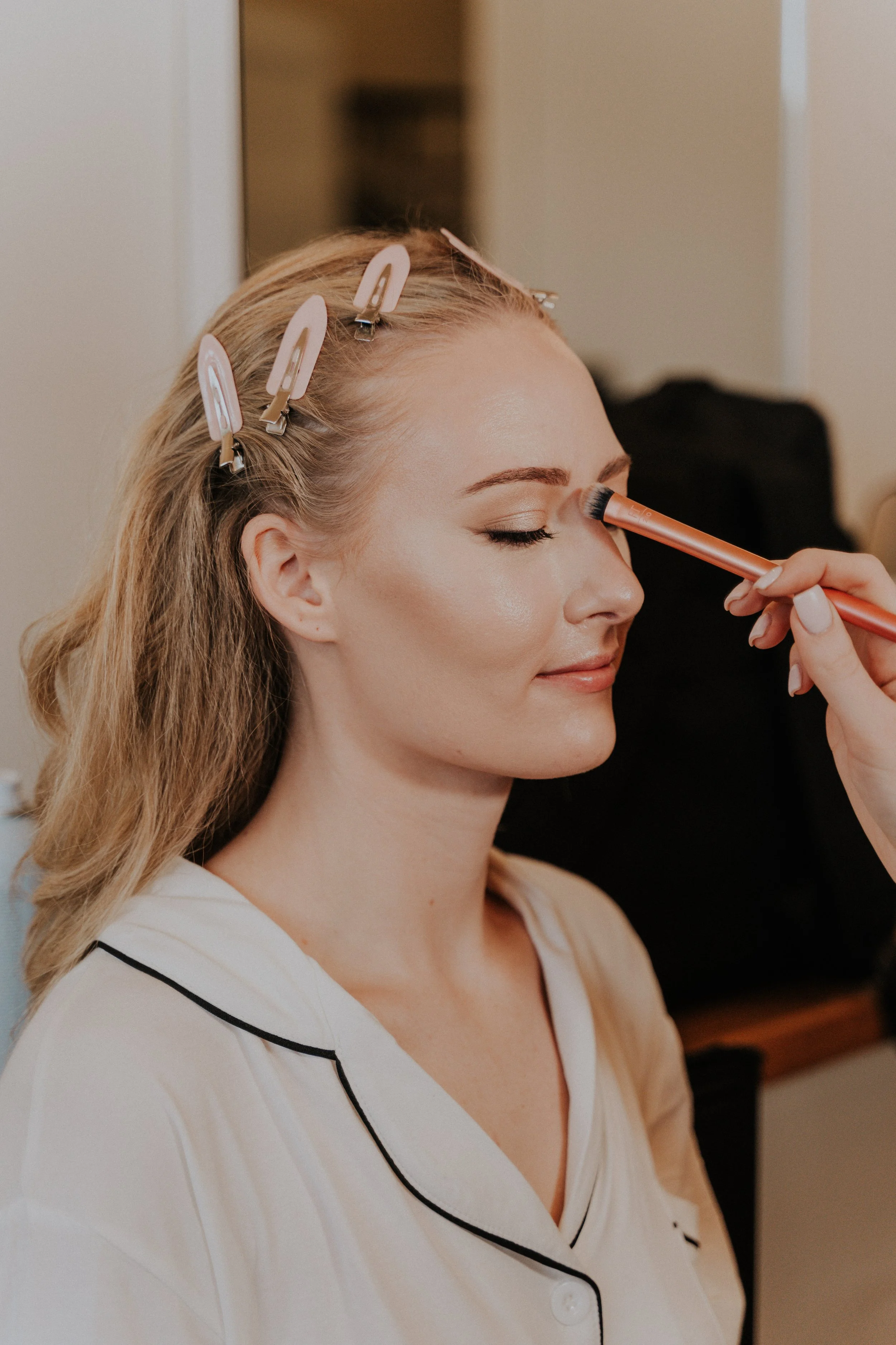 A woman with blonde hair secured with pink hair clips is getting her makeup done, with her eyes closed, while a makeup brush is applied to her eyelid.