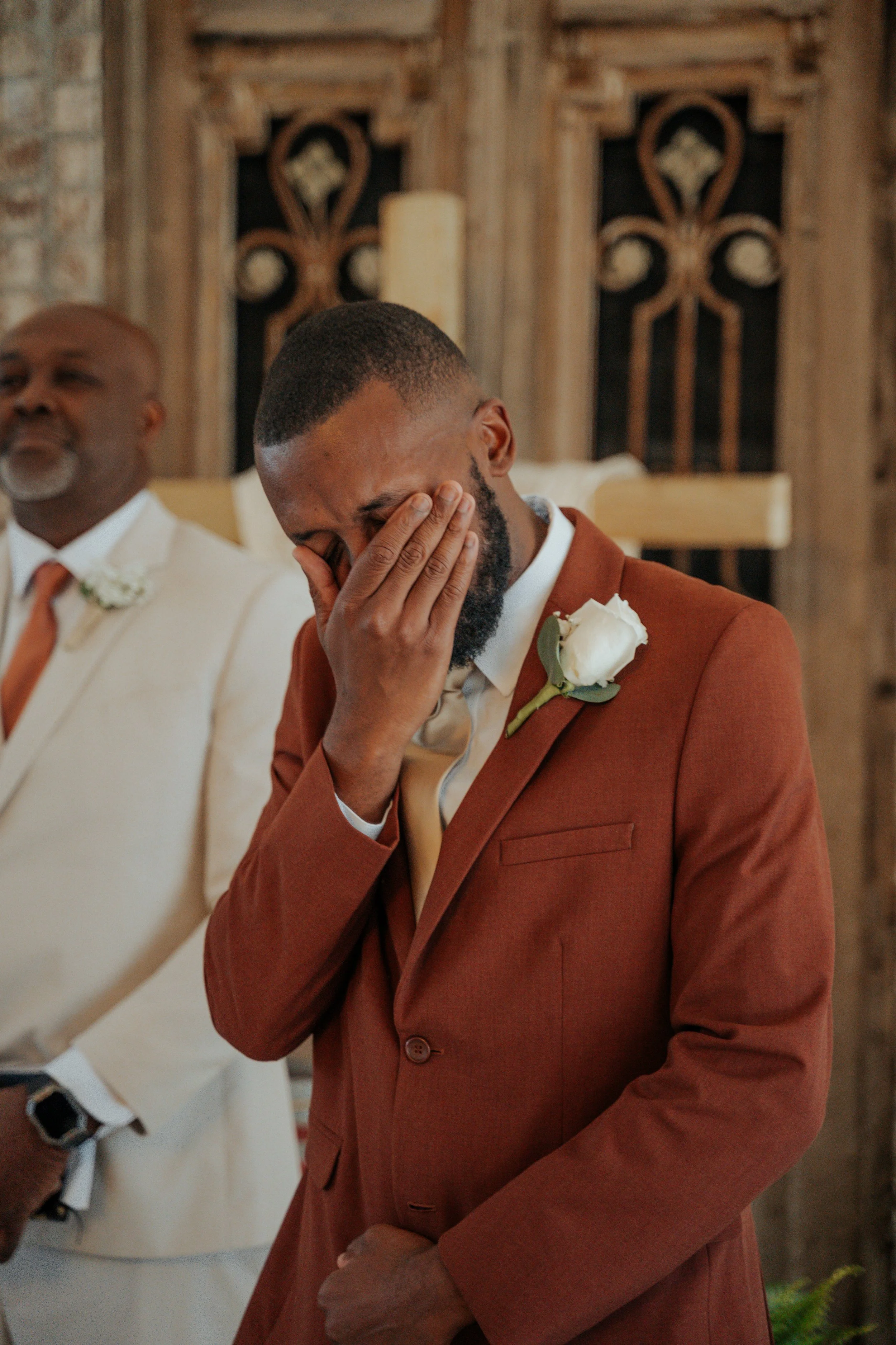 A man in a rust-colored suit with a white boutonniere covering his face with his hand, standing next to an older man in a cream-colored suit at a wedding or formal event in a rustic venue.