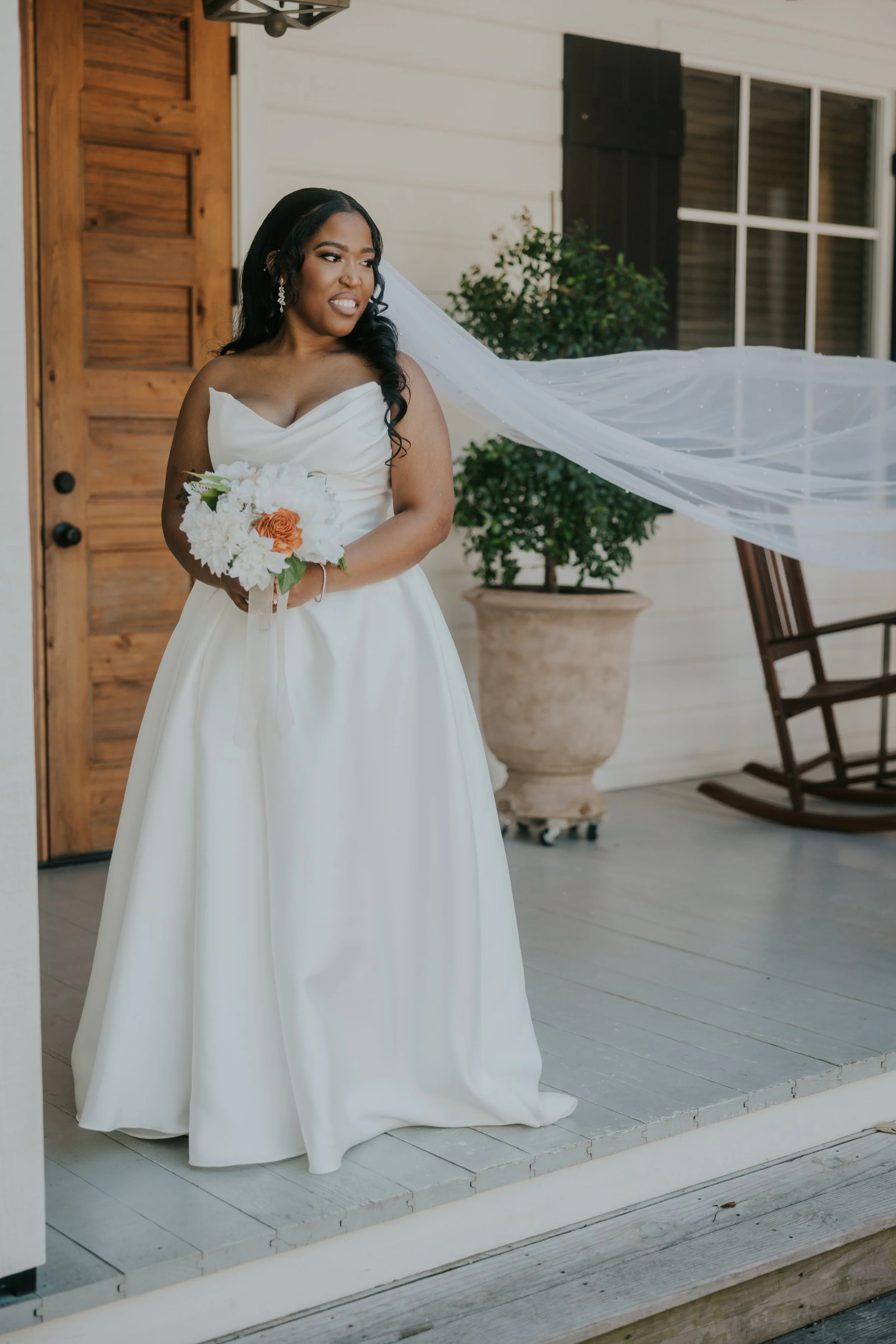 A bride in a white wedding dress holding a bouquet of flowers, standing on a porch with a rocking chair and potted plant in the background.