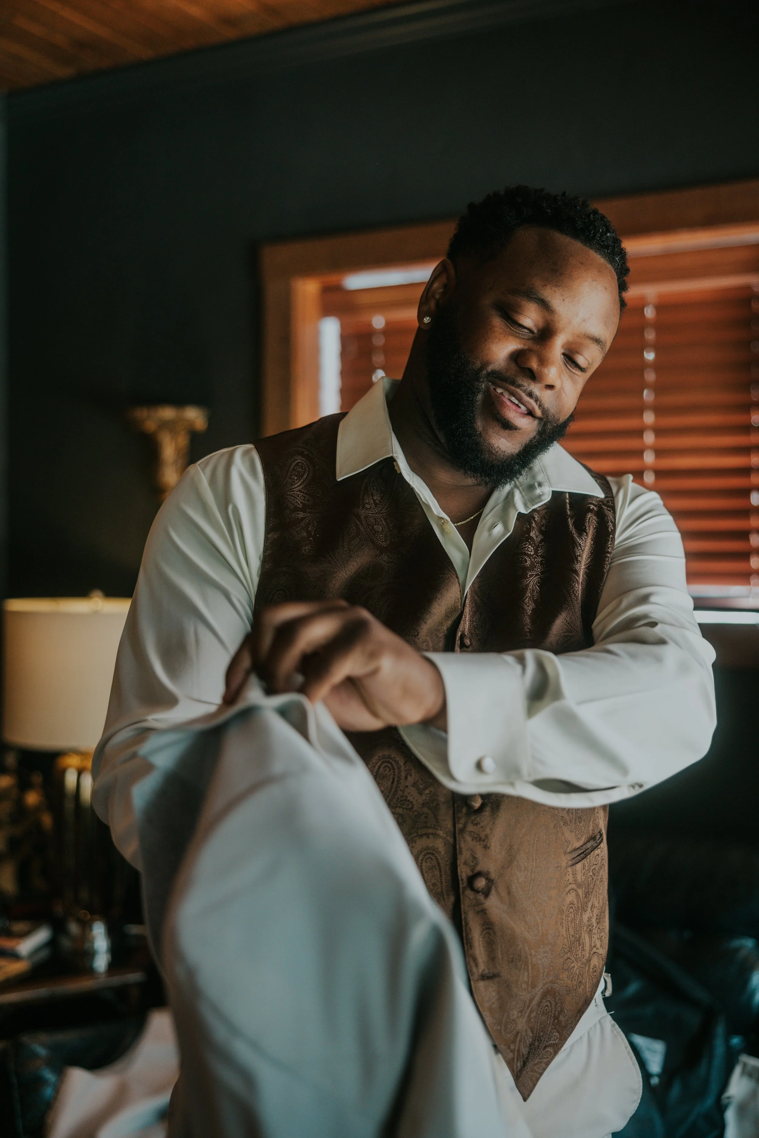 A man in a white shirt and brown vest adjusting a suit jacket, smiling indoors with a window with blinds behind him.
