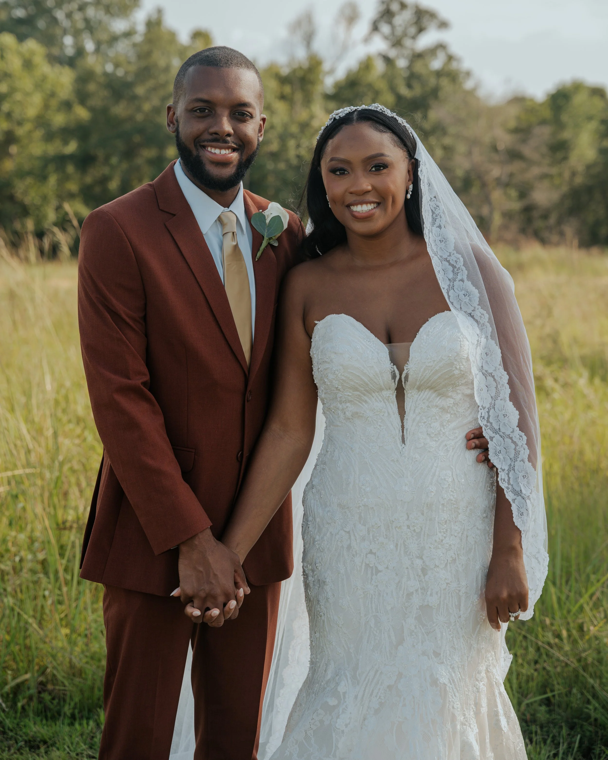 A happy couple dressed in wedding attire standing outdoors, holding hands, with green trees and grass in the background.