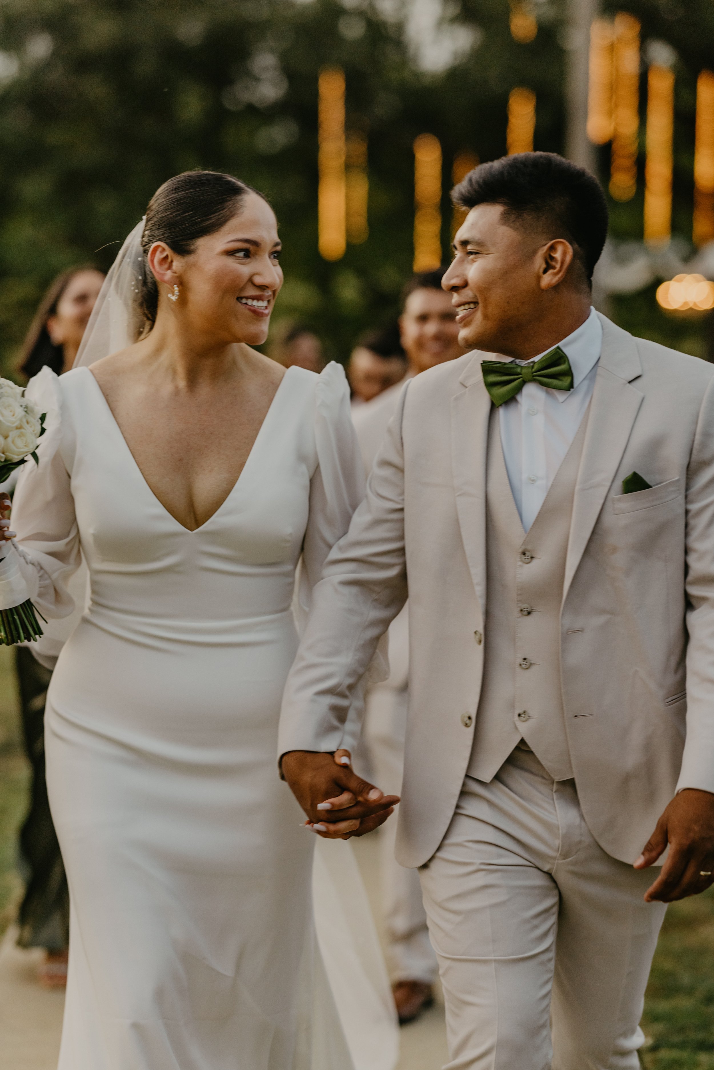 A bride and groom holding hands, smiling at each other during their wedding ceremony outdoors.