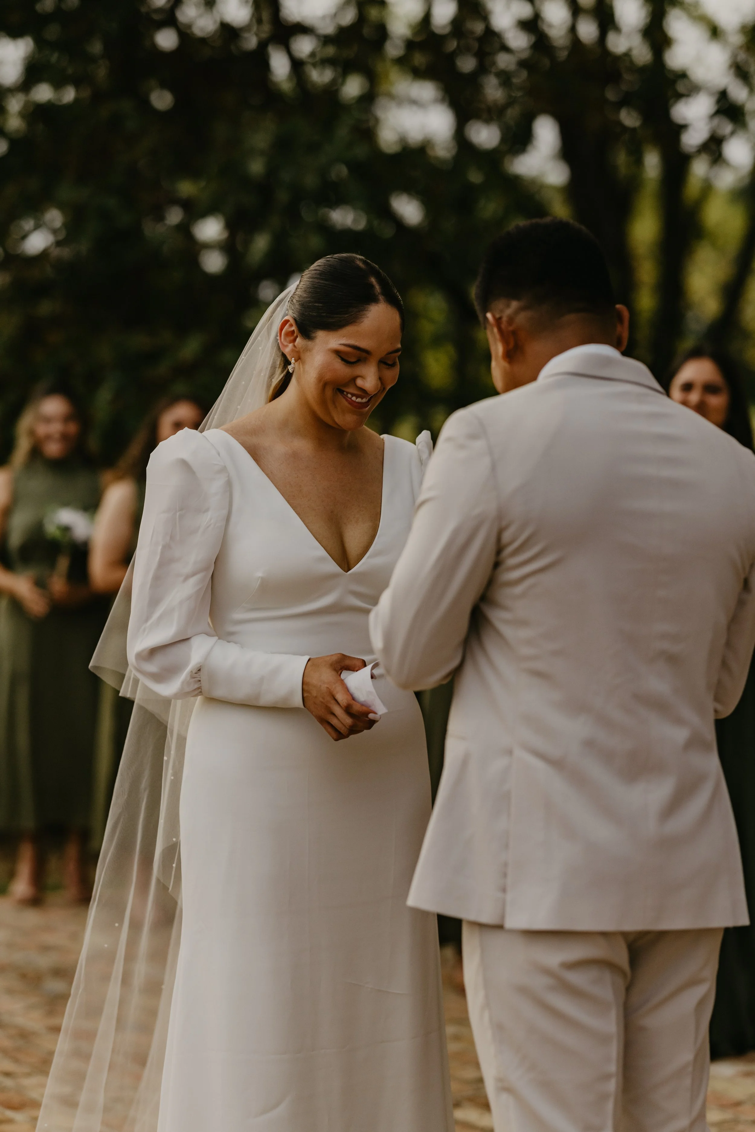 A woman and man exchanging wedding vows outdoors, with women standing behind them, blurred in the background.