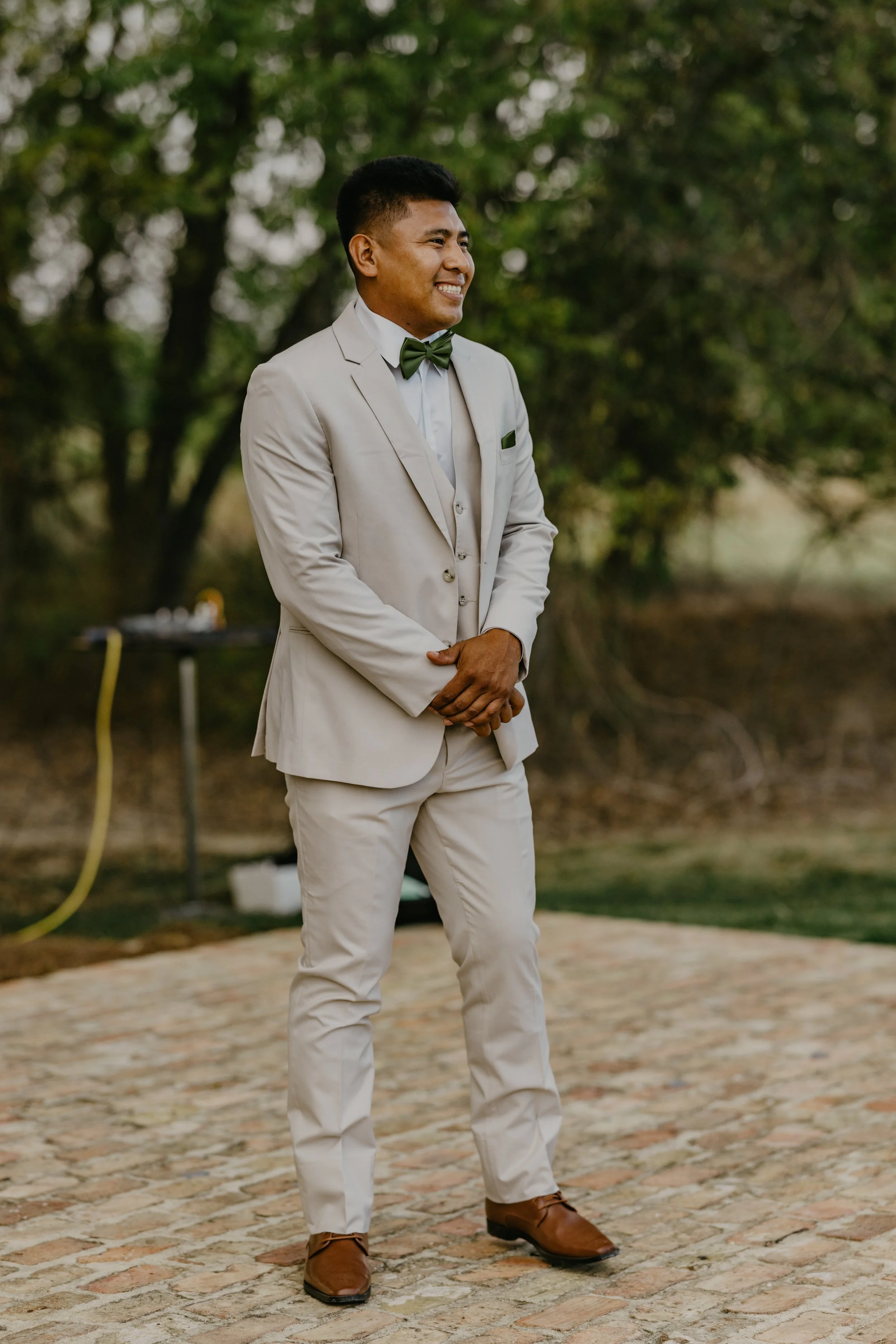 A man dressed in a light beige suit with a matching vest, white shirt, green bow tie, and brown shoes stands outdoors on a cobblestone surface during daytime. He is smiling and standing in front of green trees.