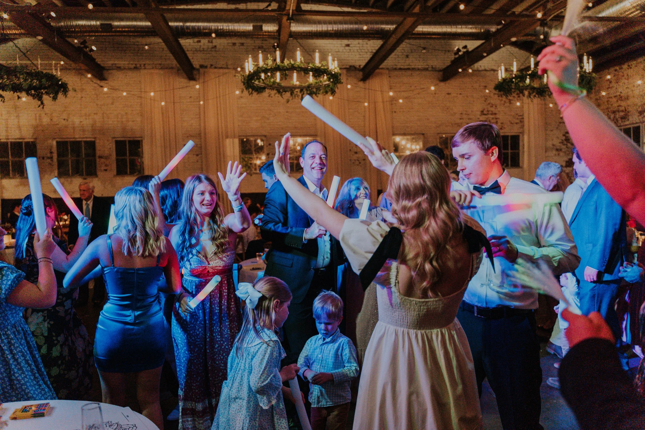 People celebrating at a party with glowing sticks, dancing, and smiling in a warmly lit venue with hanging lights and wreath decorations.