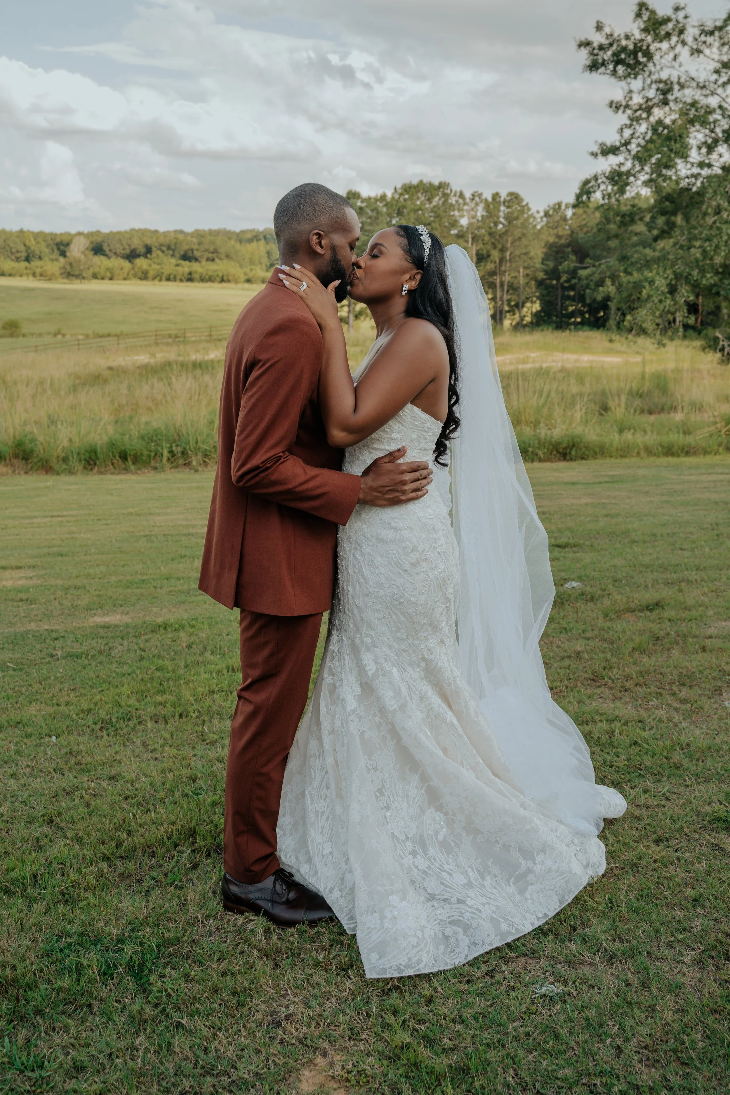 A bride and groom sharing a kiss outdoors in a grassy field with trees in the background. The bride wears a white lace wedding gown and a veil, and the groom wears a brown suit.