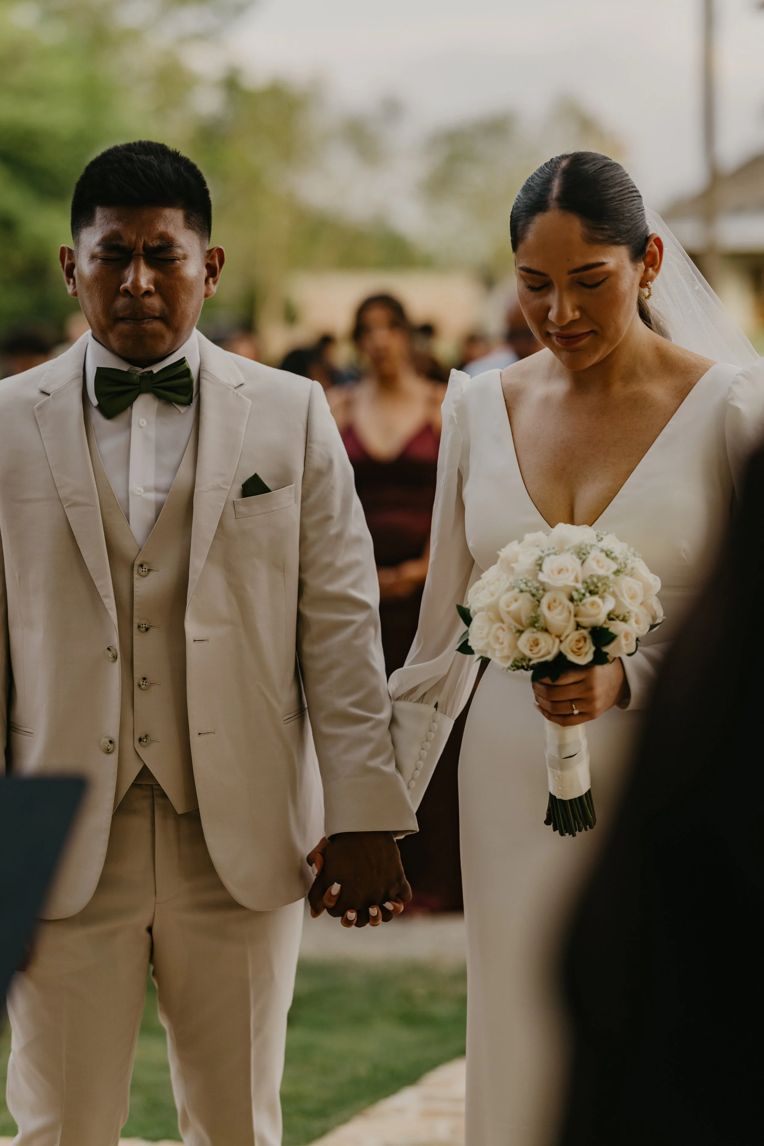 A bride and groom holding hands during their wedding ceremony, with the bride holding a bouquet of white roses and the groom dressed in a beige suit with a green bowtie.