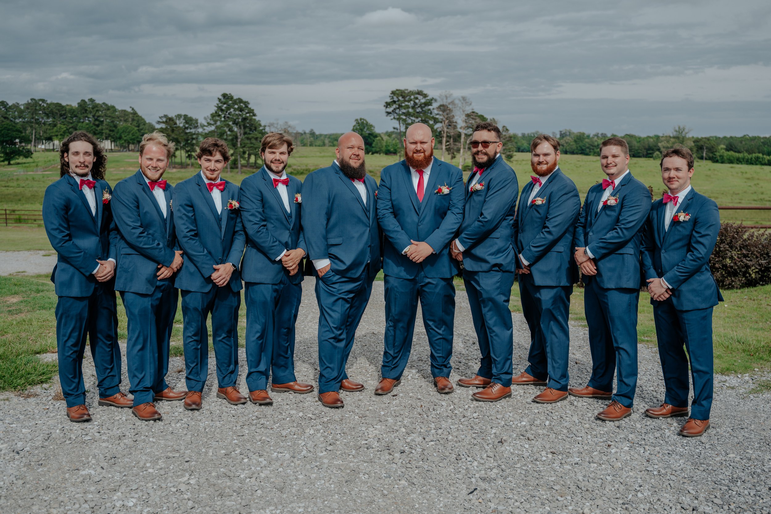 A group of eleven men dressed in matching blue suits and red bow ties, standing outdoors on a gravel path with green fields and trees in the background, during a wedding or formal event.