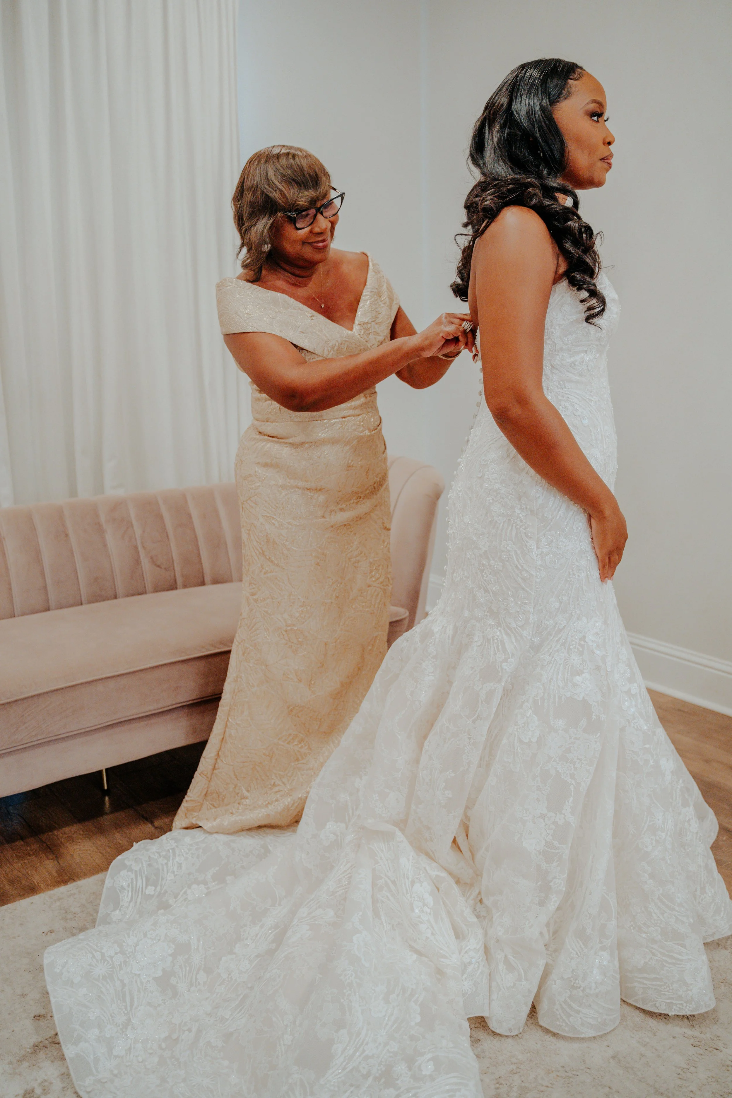 A woman in a white wedding dress standing on a beige carpet while another woman in a gold dress adjusts her back.