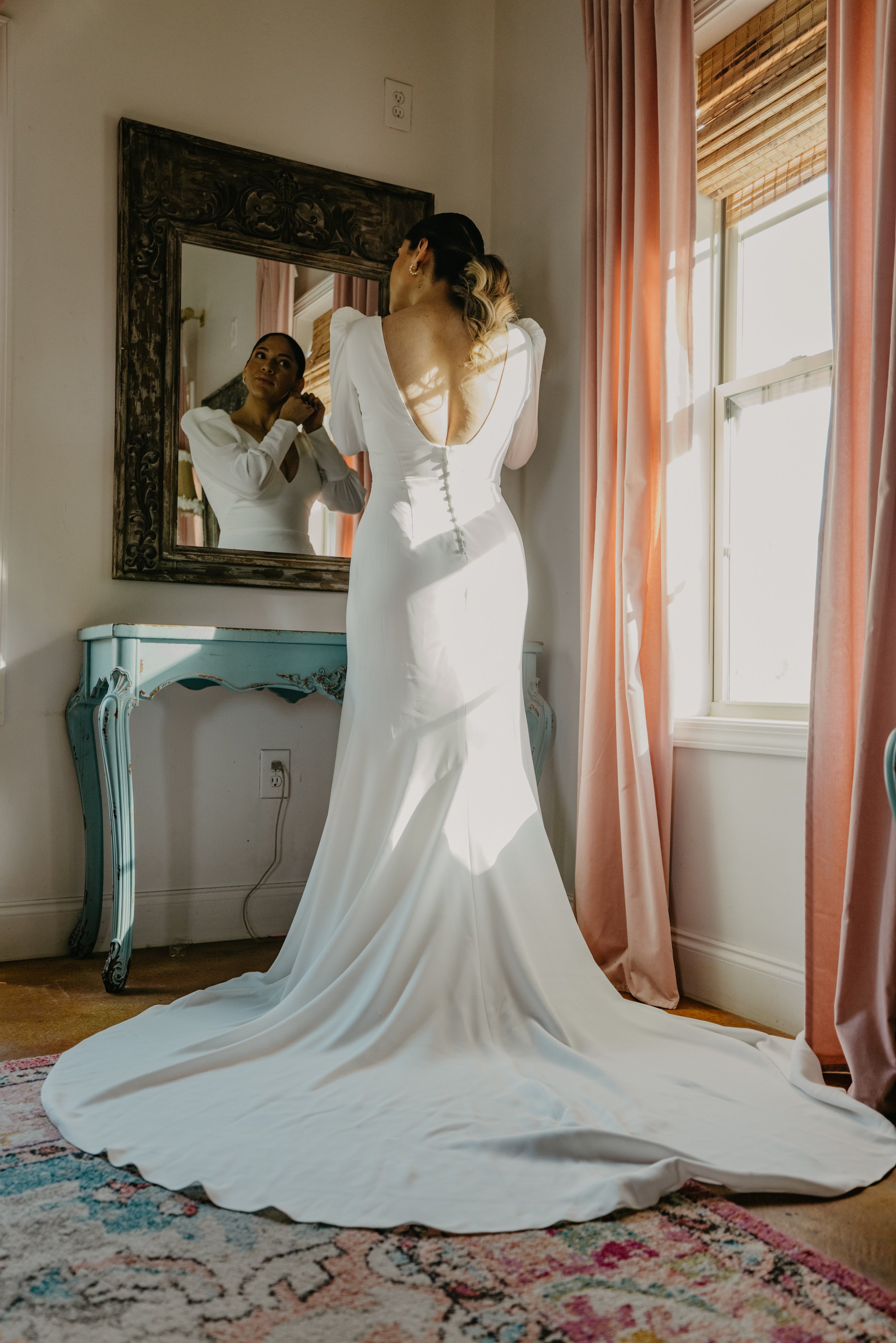 A bride in a white wedding gown adjusting her earrings in front of a mirror, standing next to a window with pink curtains, in a room with a vintage blue table and patterned rug.
