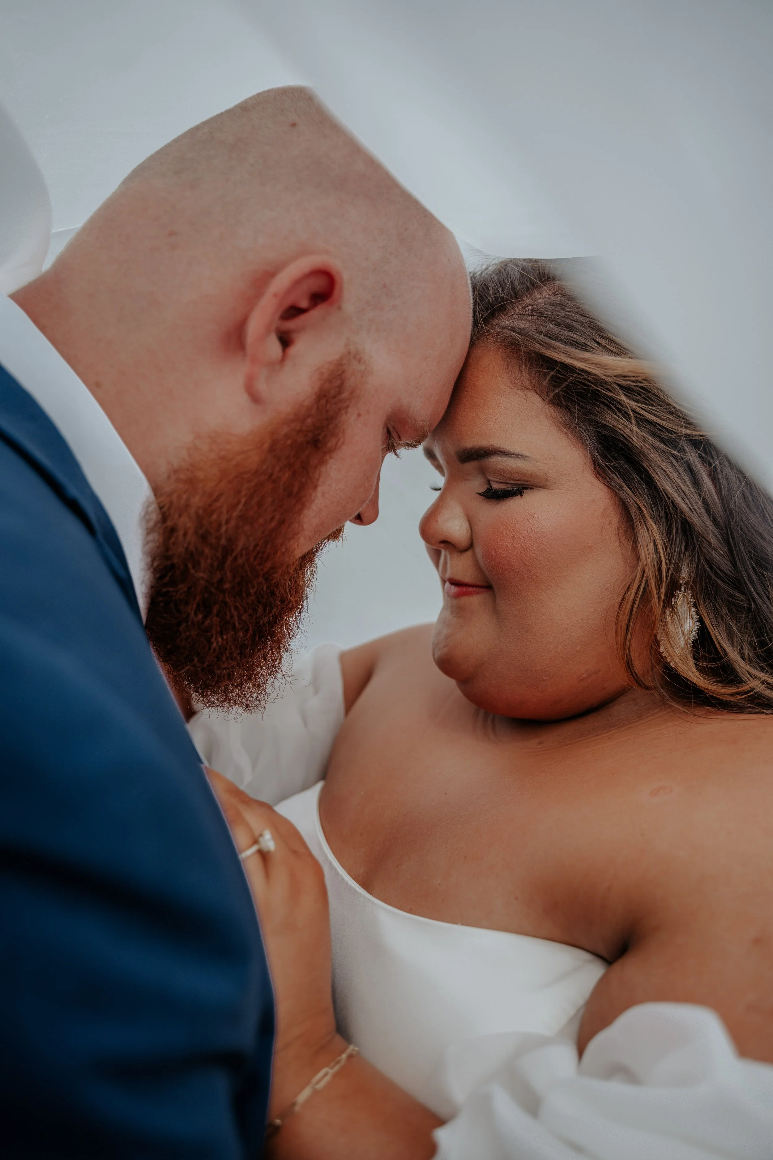 A close-up of a tender moment between a newlywed couple with their foreheads touching, eyes closed, inside a white wedding venue.