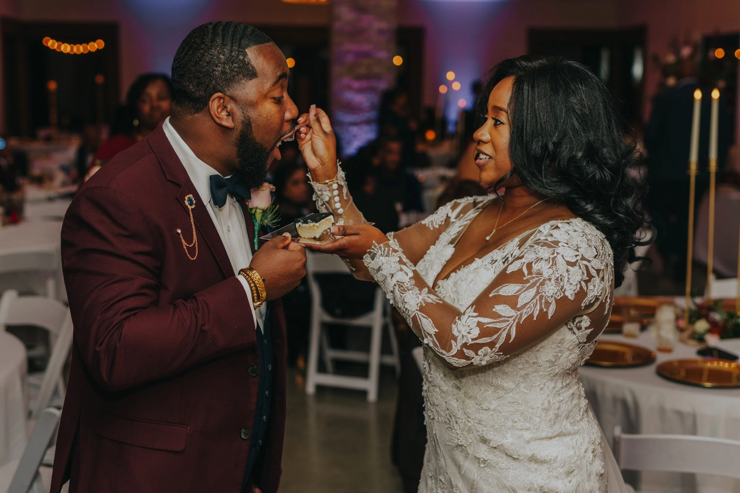 A bride in a lace wedding dress feeds cake to a groom in a burgundy suit at a wedding reception.