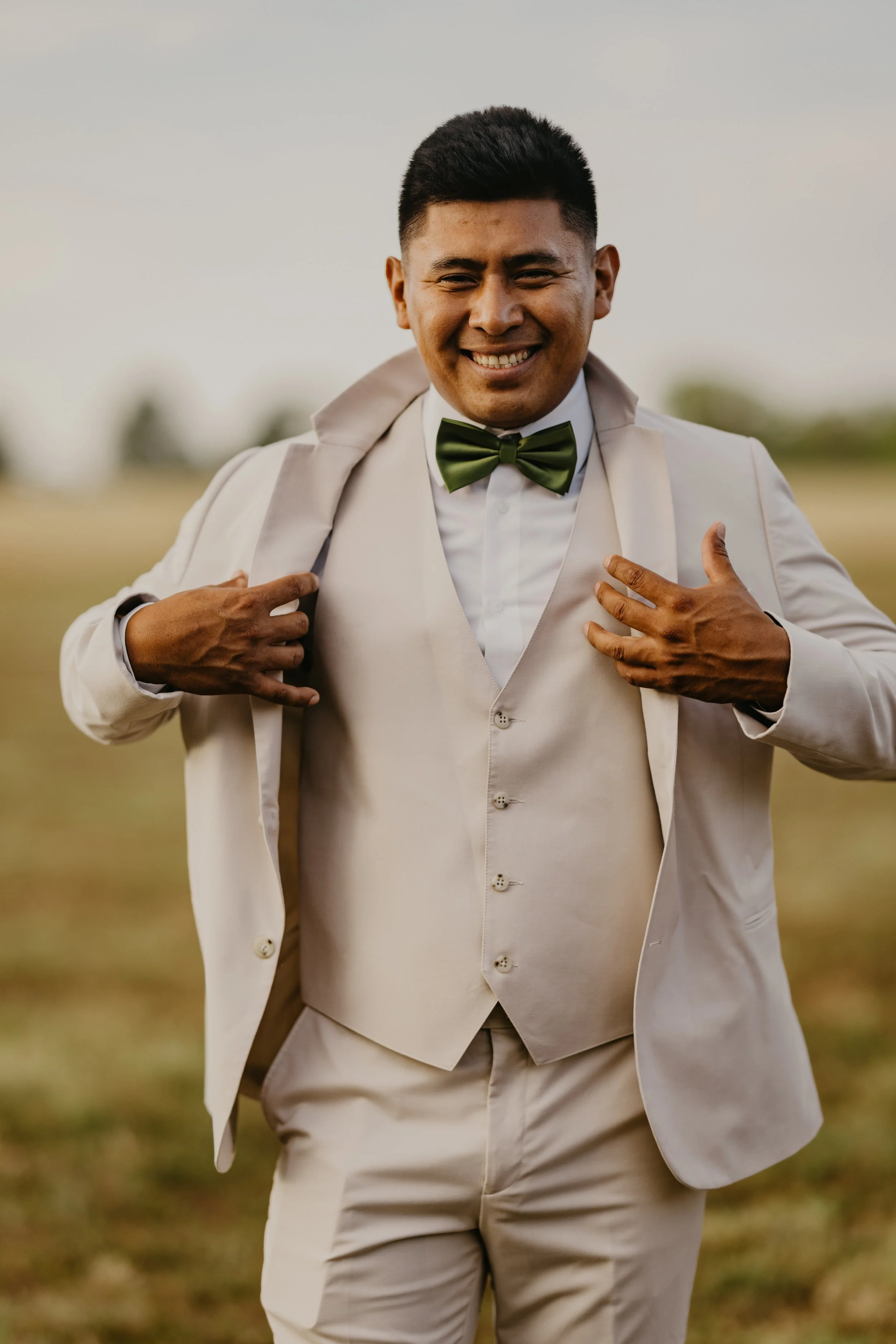 A smiling man in a beige suit, green bow tie, and white shirt standing outdoors in a field with his jacket draped over his shoulders.