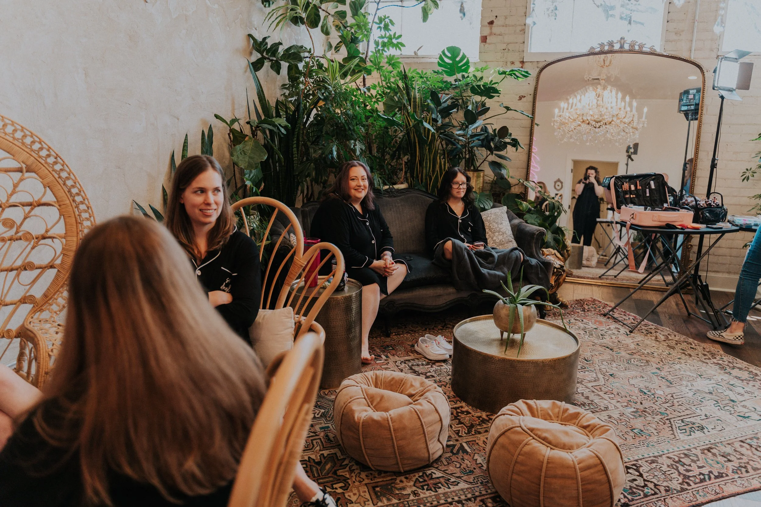 Women seated on a black sofa and wicker chairs in a cozy indoor space, with lush green plants behind them and a large mirror reflecting a chandelier and a person taking a photo, surrounded by makeup and photography equipment.