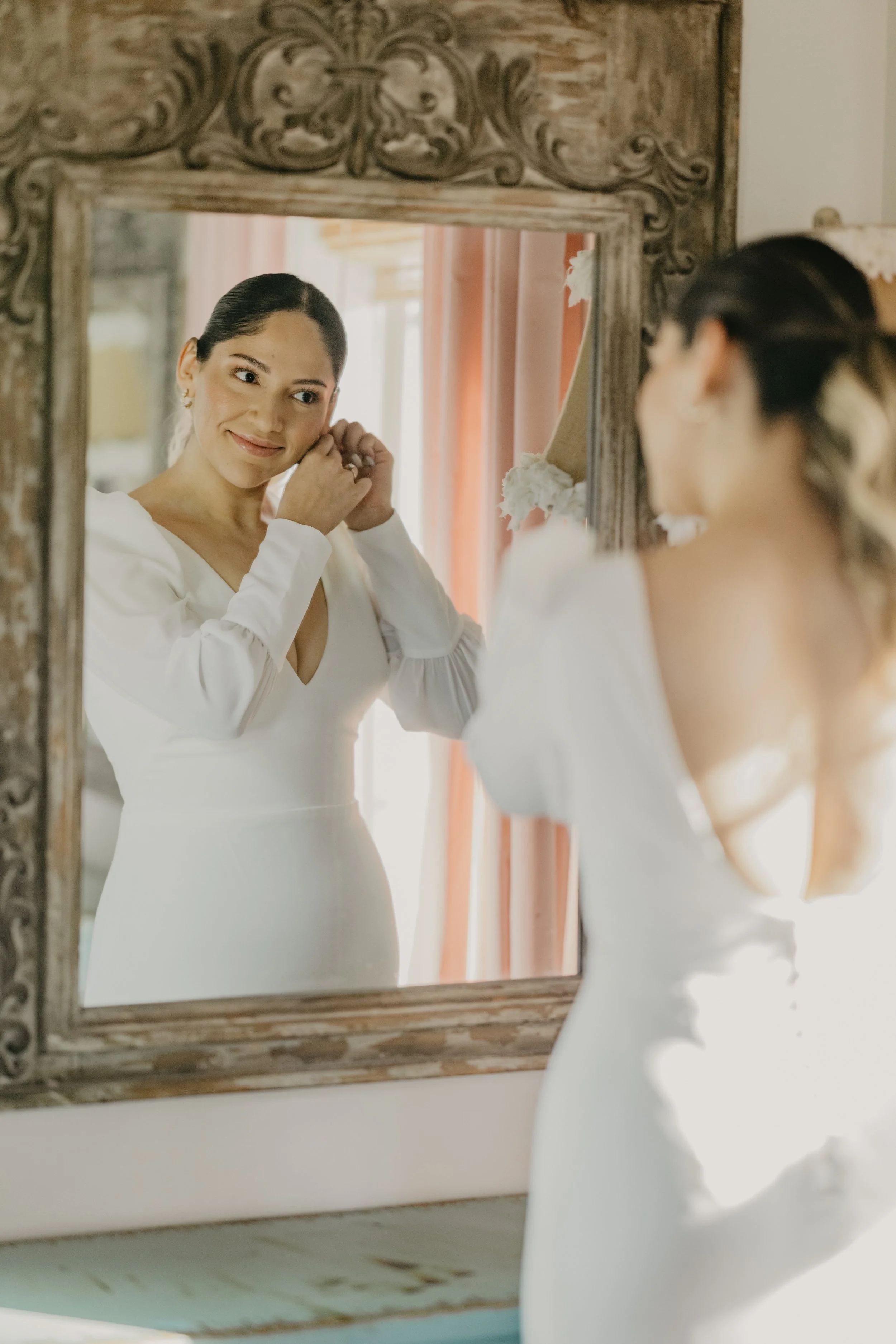 A bride in a white dress is looking at her reflection in a mirror while adjusting her earrings.