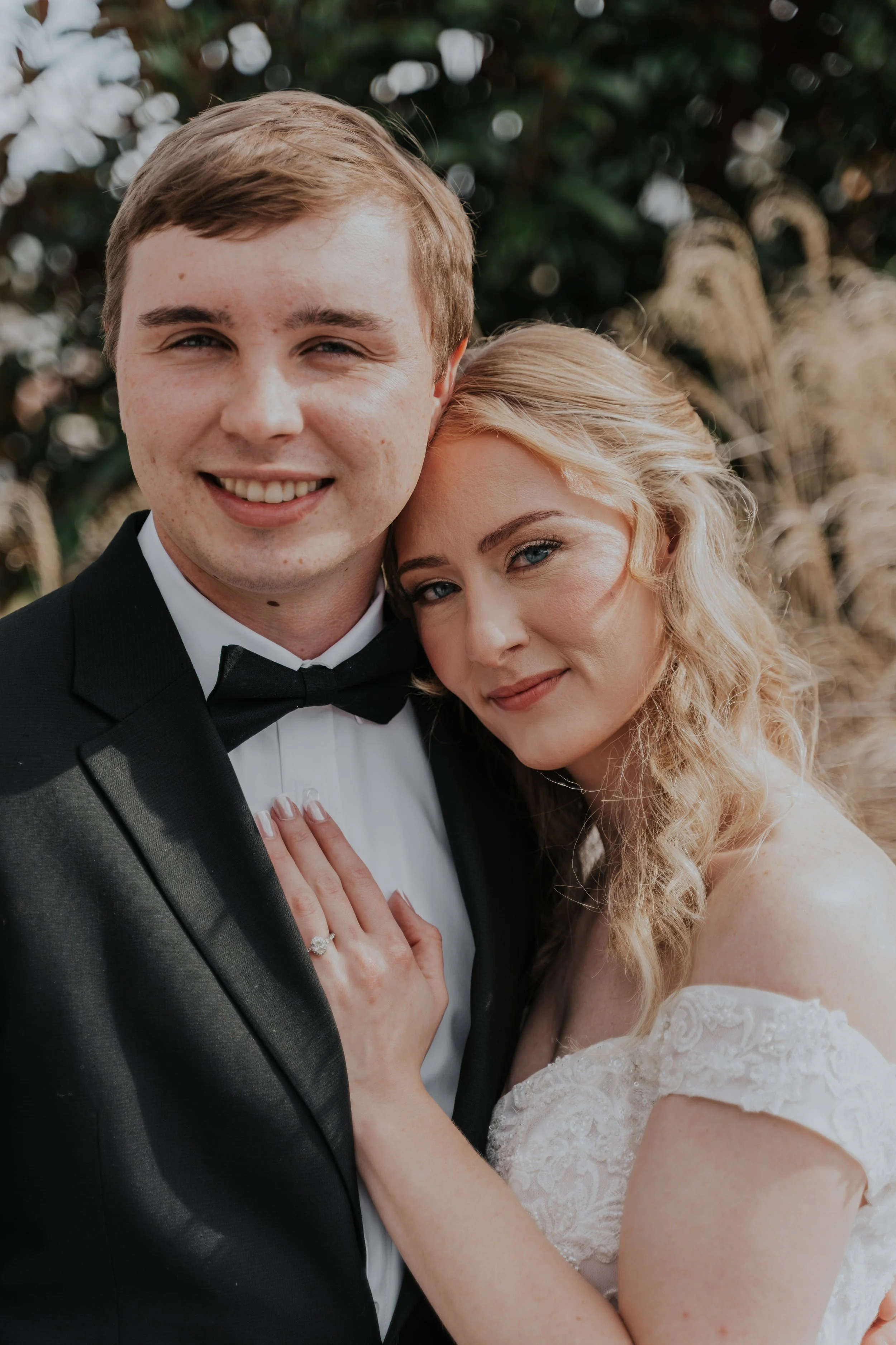 A happy bride and groom pose closely in wedding attire outdoors, with the bride showing her wedding ring.