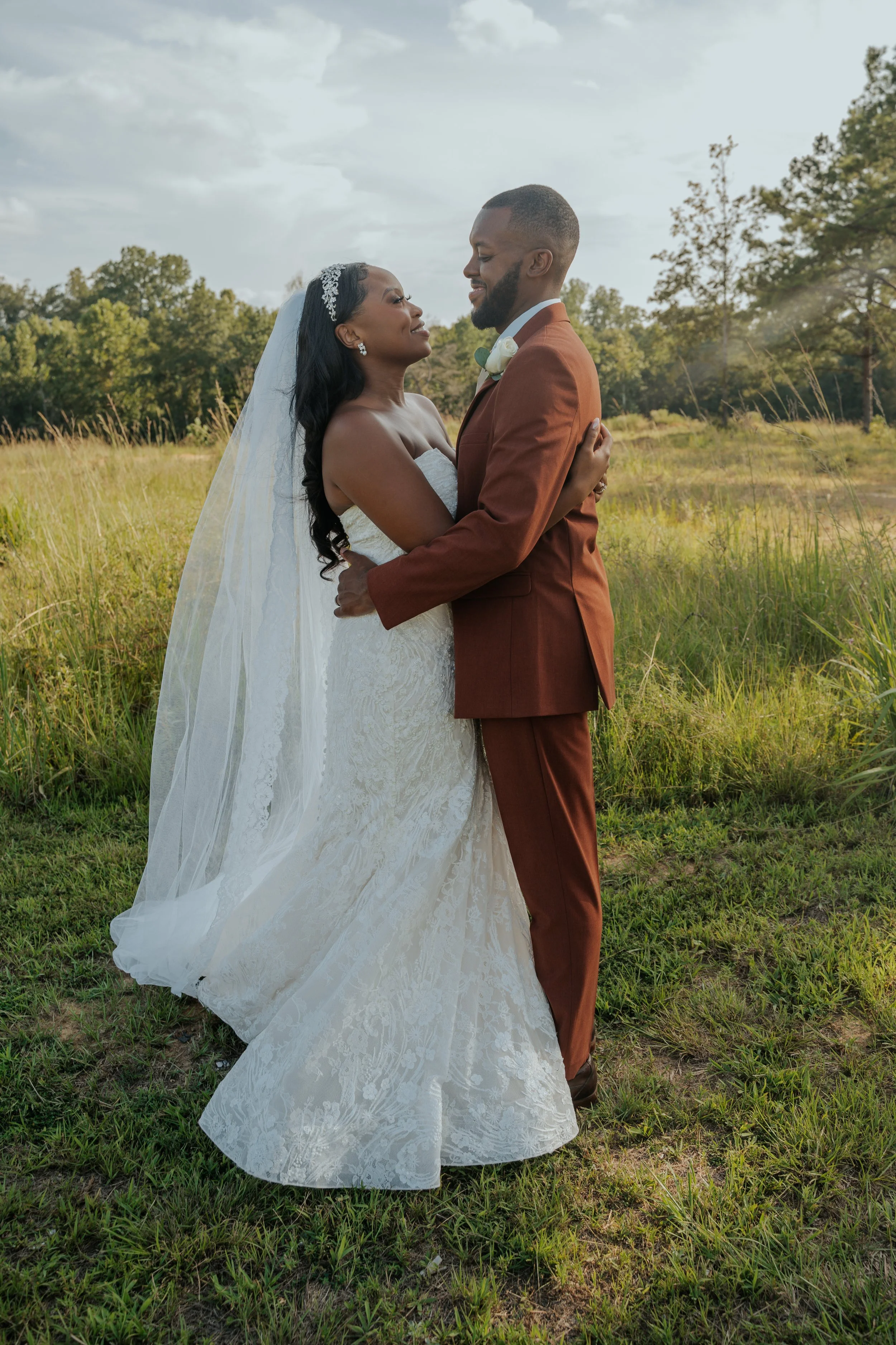 A bride and groom in wedding attire embracing outdoors in a grassy field, with trees and a cloudy sky in the background.