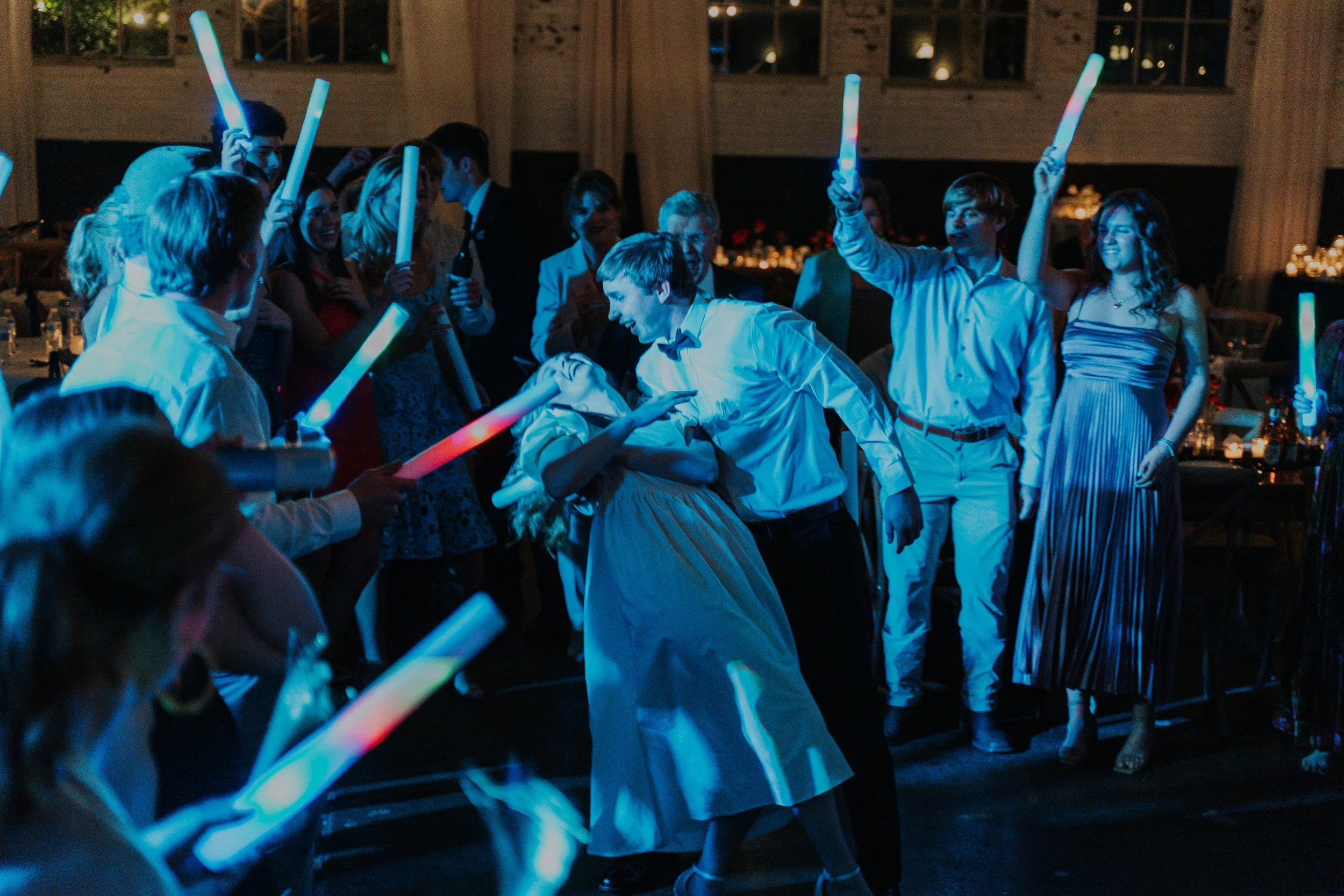 People dancing and celebrating at a party with glow sticks in a dimly lit room.