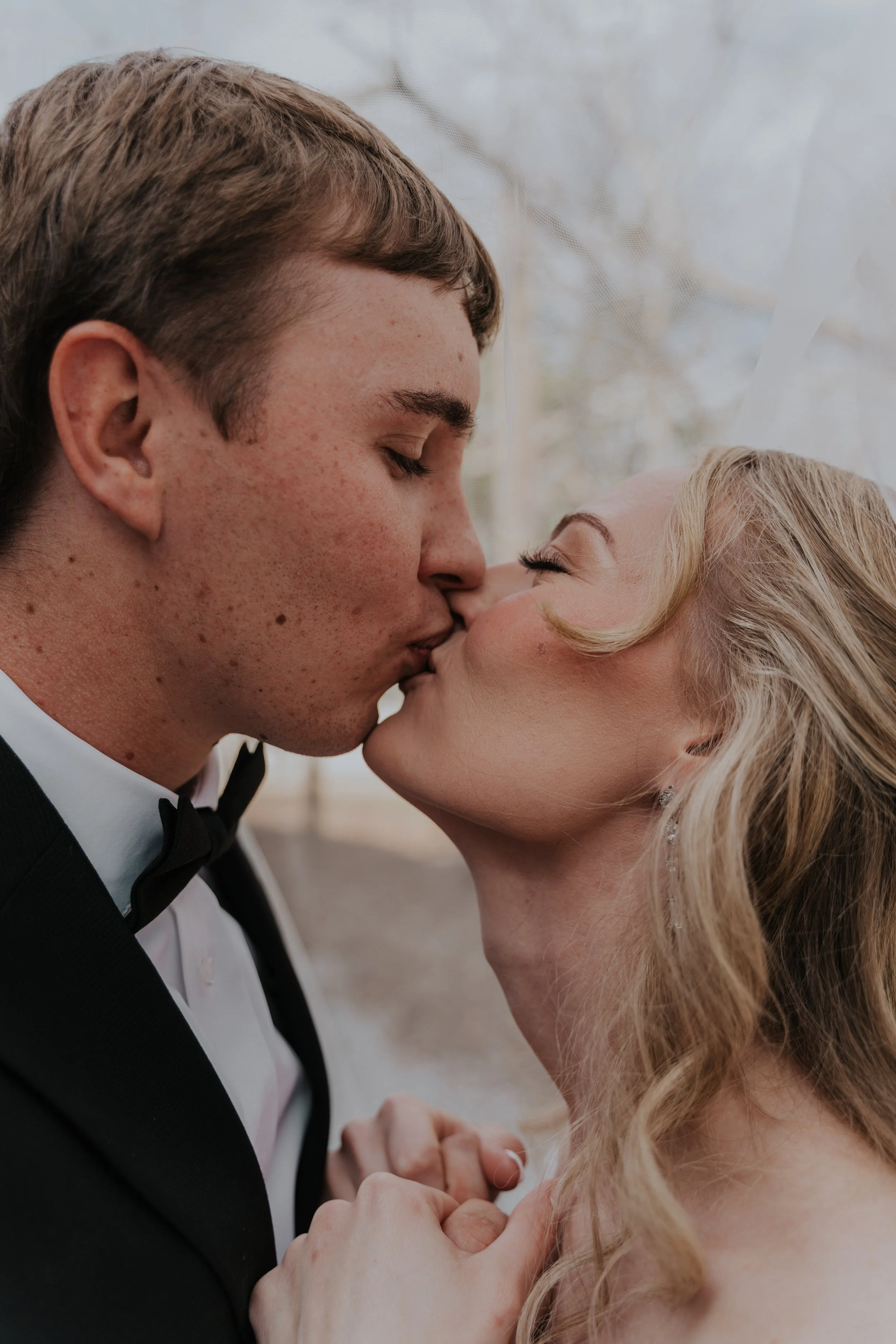 A bride and groom sharing a kiss, with the groom in a tuxedo and bowtie and the bride with blonde hair and earrings, holding hands.