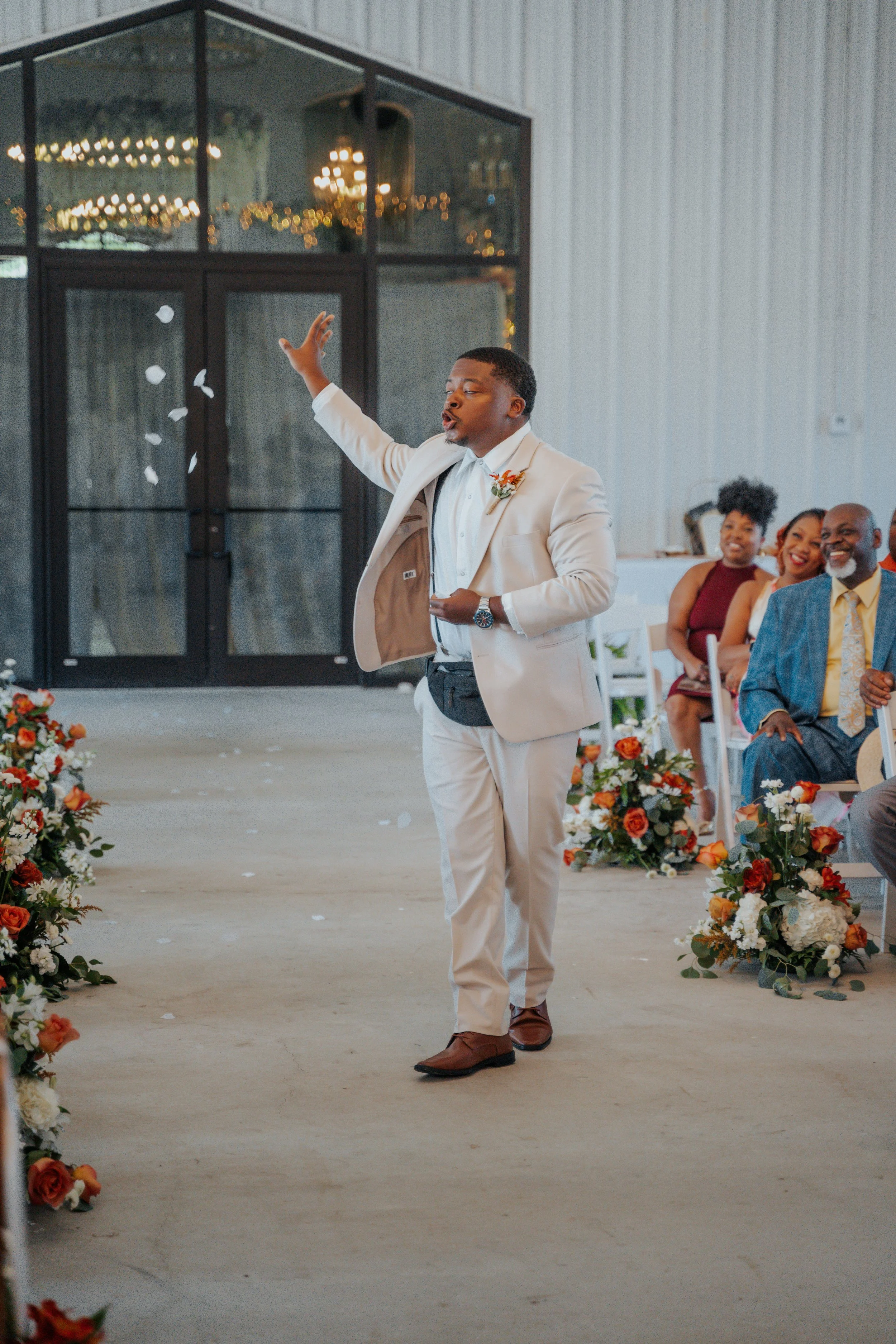 A man in a cream suit stands on a decorated aisle at a wedding, tossing flower petals, with guests seated on either side, smiling and watching.