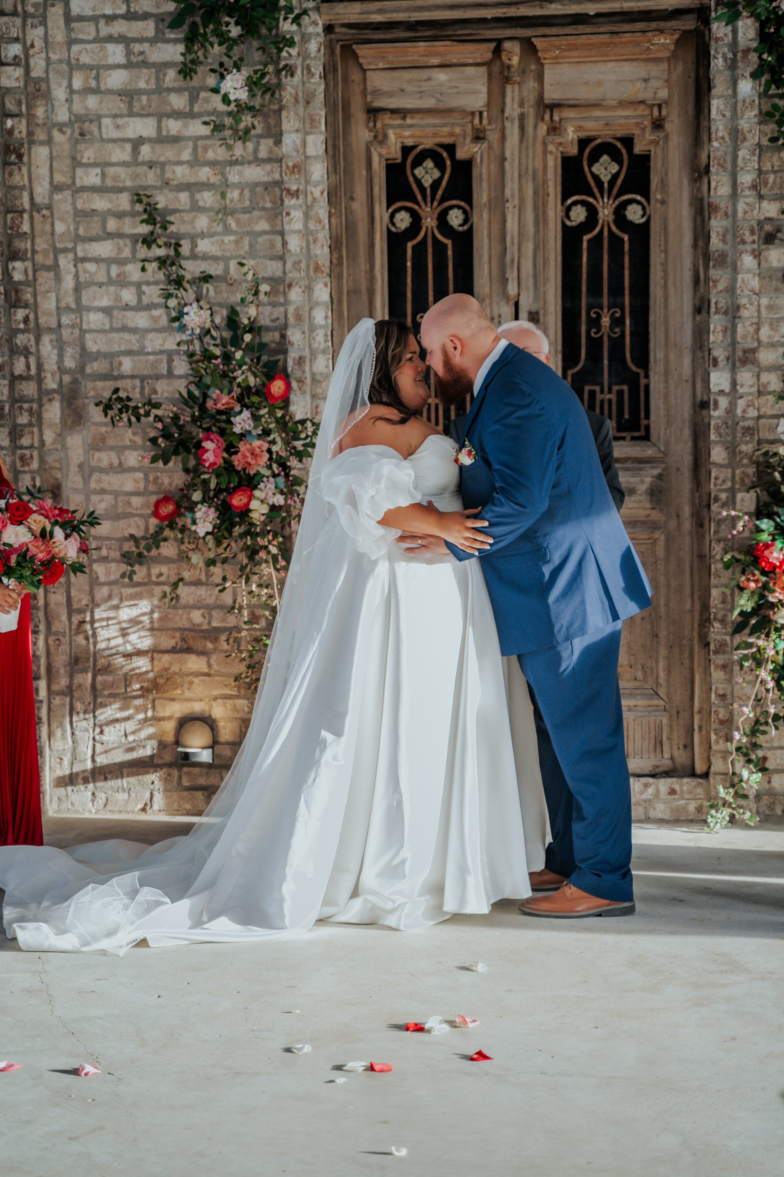 A bride and groom sharing a kiss during their wedding ceremony, decorated with flowers and set against a rustic wooden door and brick wall.