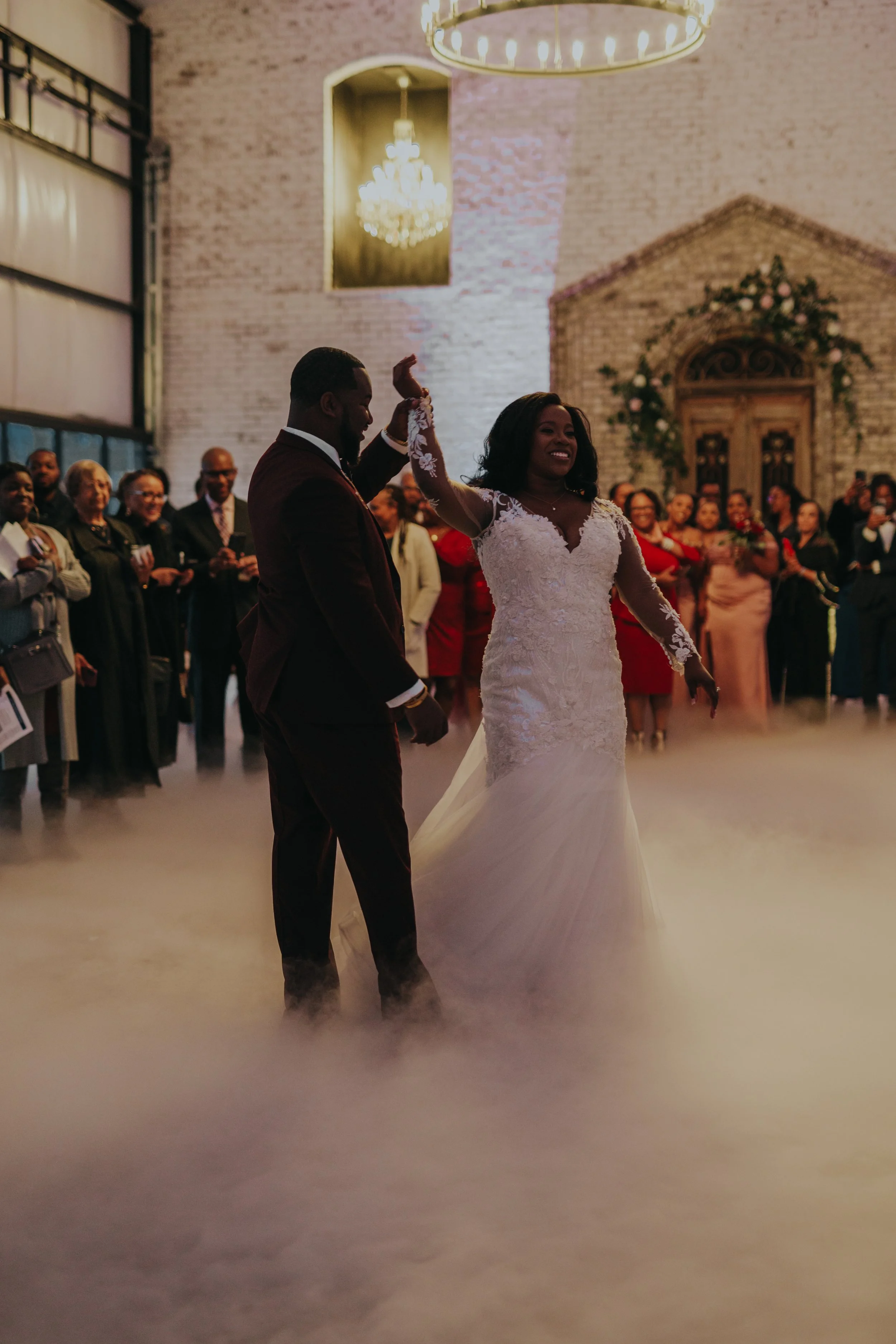 A bride and groom dancing at their wedding reception, surrounded by guests in an elegant venue with chandeliers and a brick wall.