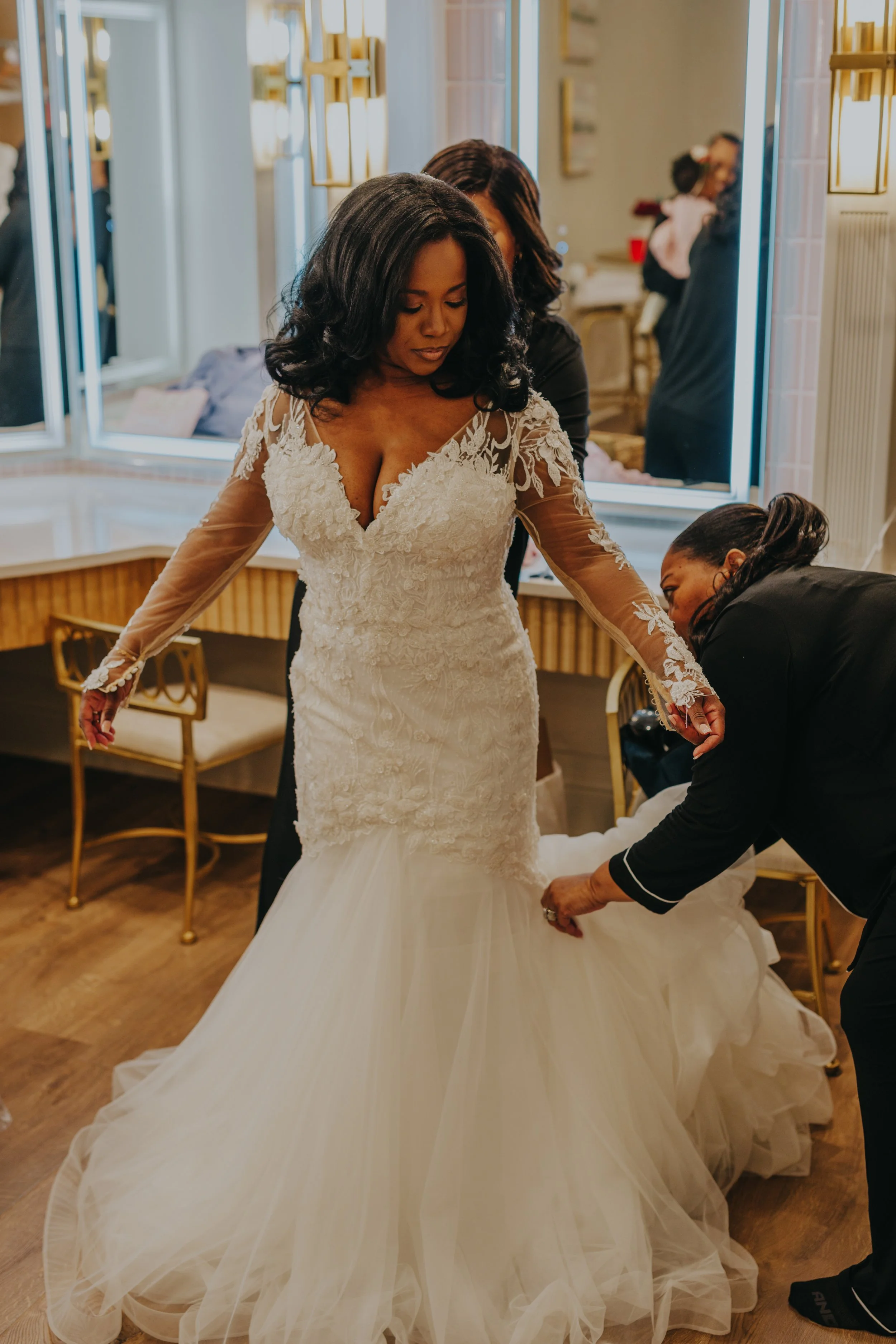 A bride in a lace and tulle wedding dress is being assisted with her gown by two women in a room with mirrors and warm lighting.