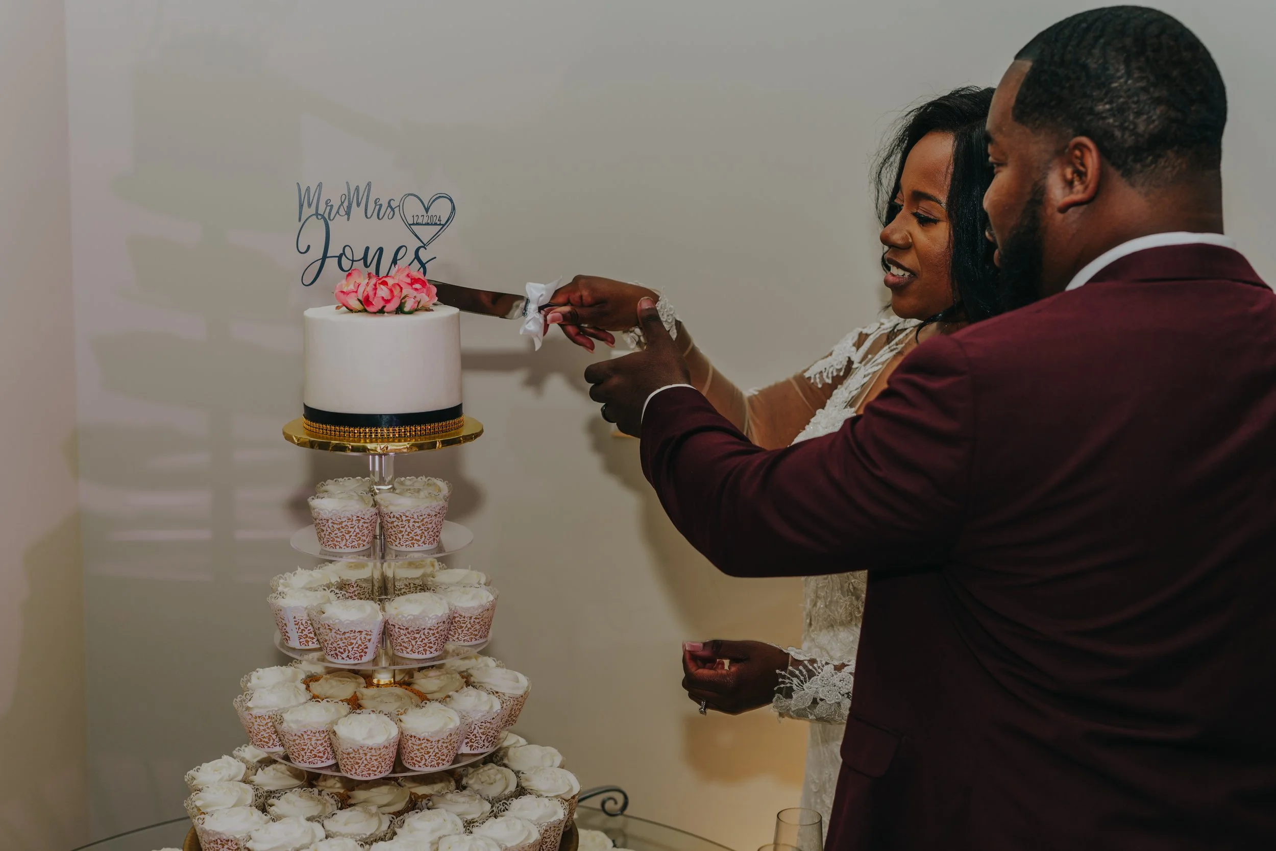 A couple is cutting a wedding cake together at their wedding reception. The cake is a multi-tiered display with cupcakes and a small, round cake on top decorated with pink flowers. The bride and groom are smiling, dressed in formal attire, with the b