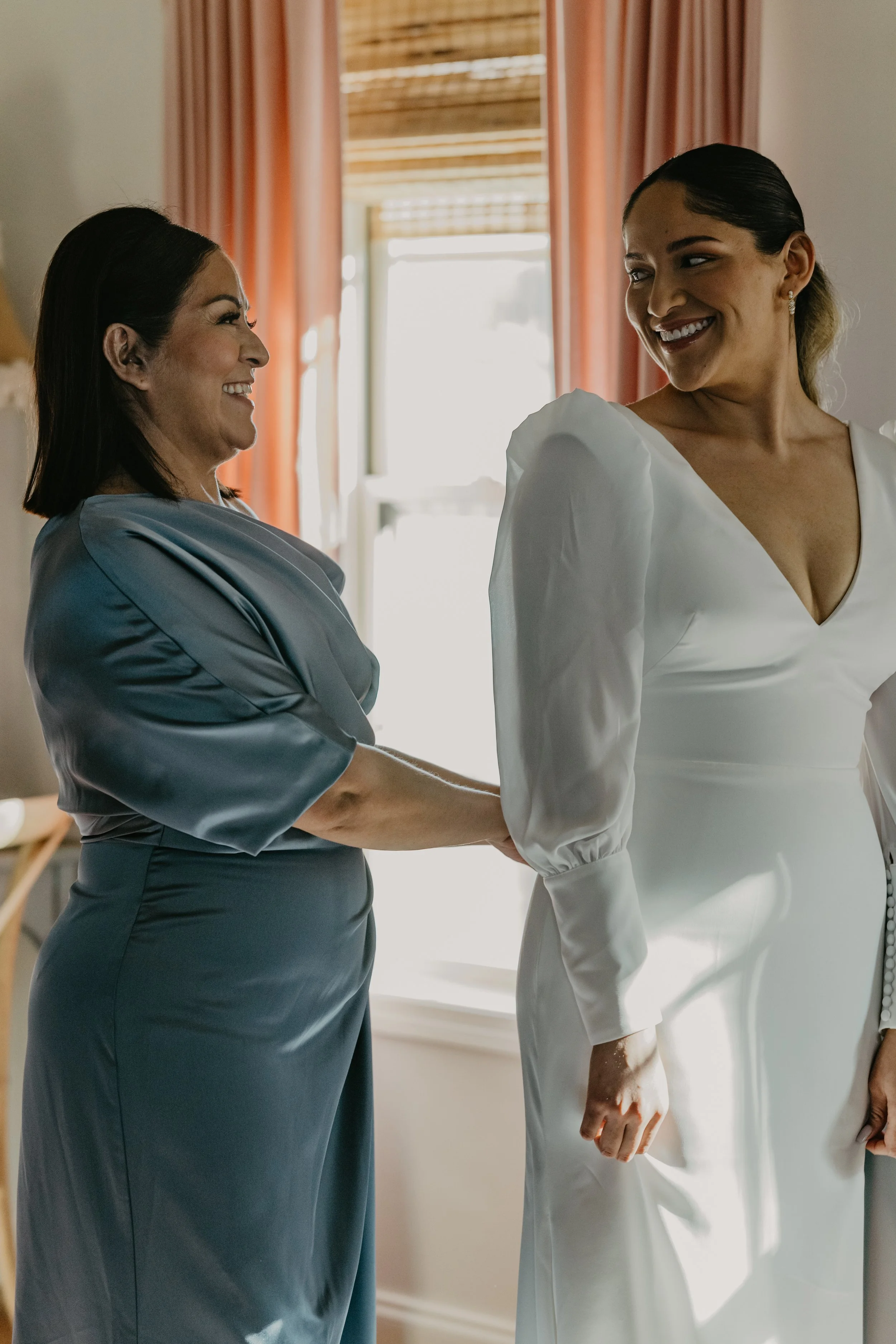 Two women smiling and holding hands, one in a gray satin dress and the other in a white wedding gown, standing indoors near a window with sunlight coming through.
