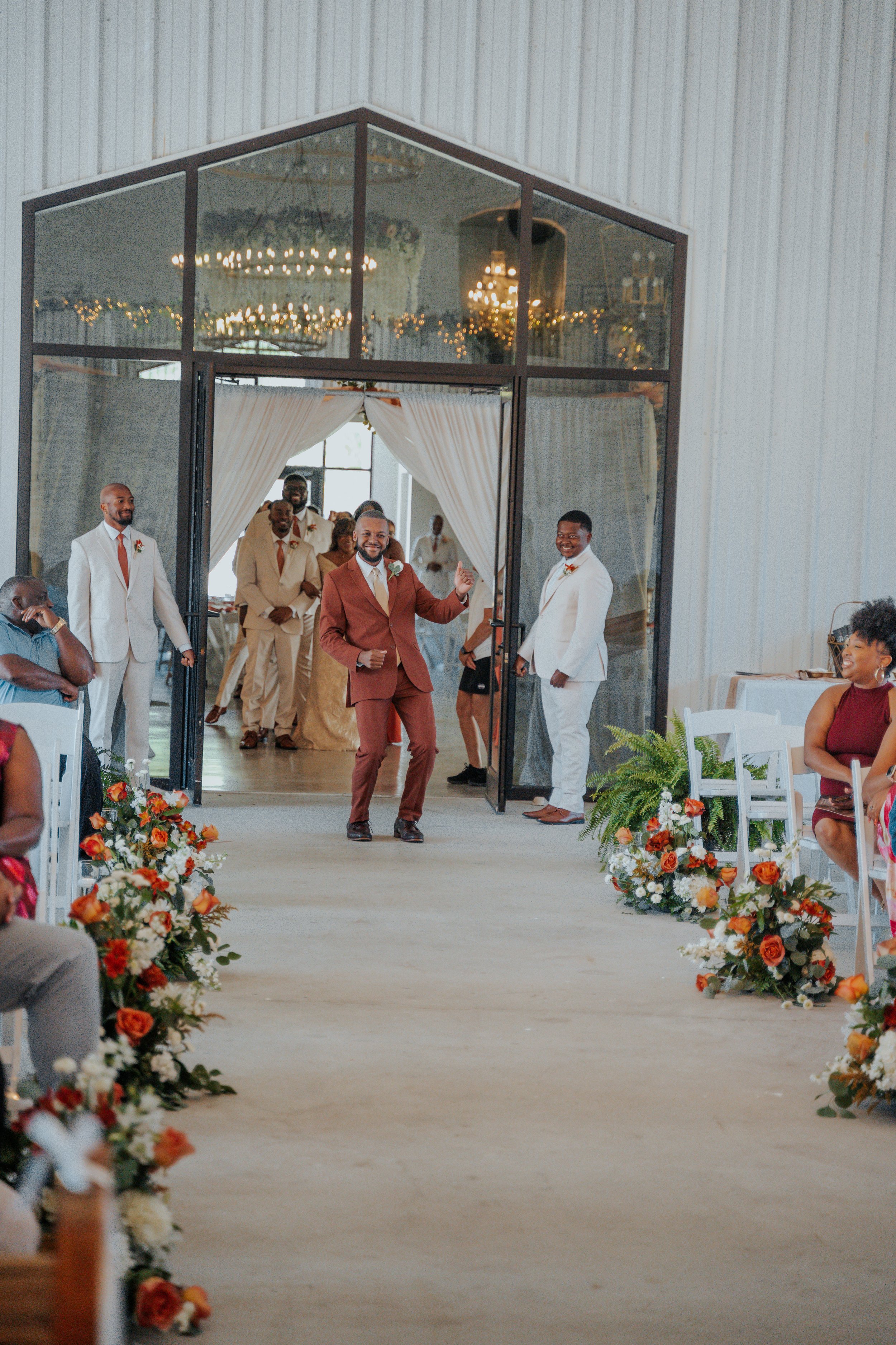 Groom walking down the aisle at a wedding ceremony, surrounded by guests, colorful floral arrangements, and a decorated venue with chandeliers.