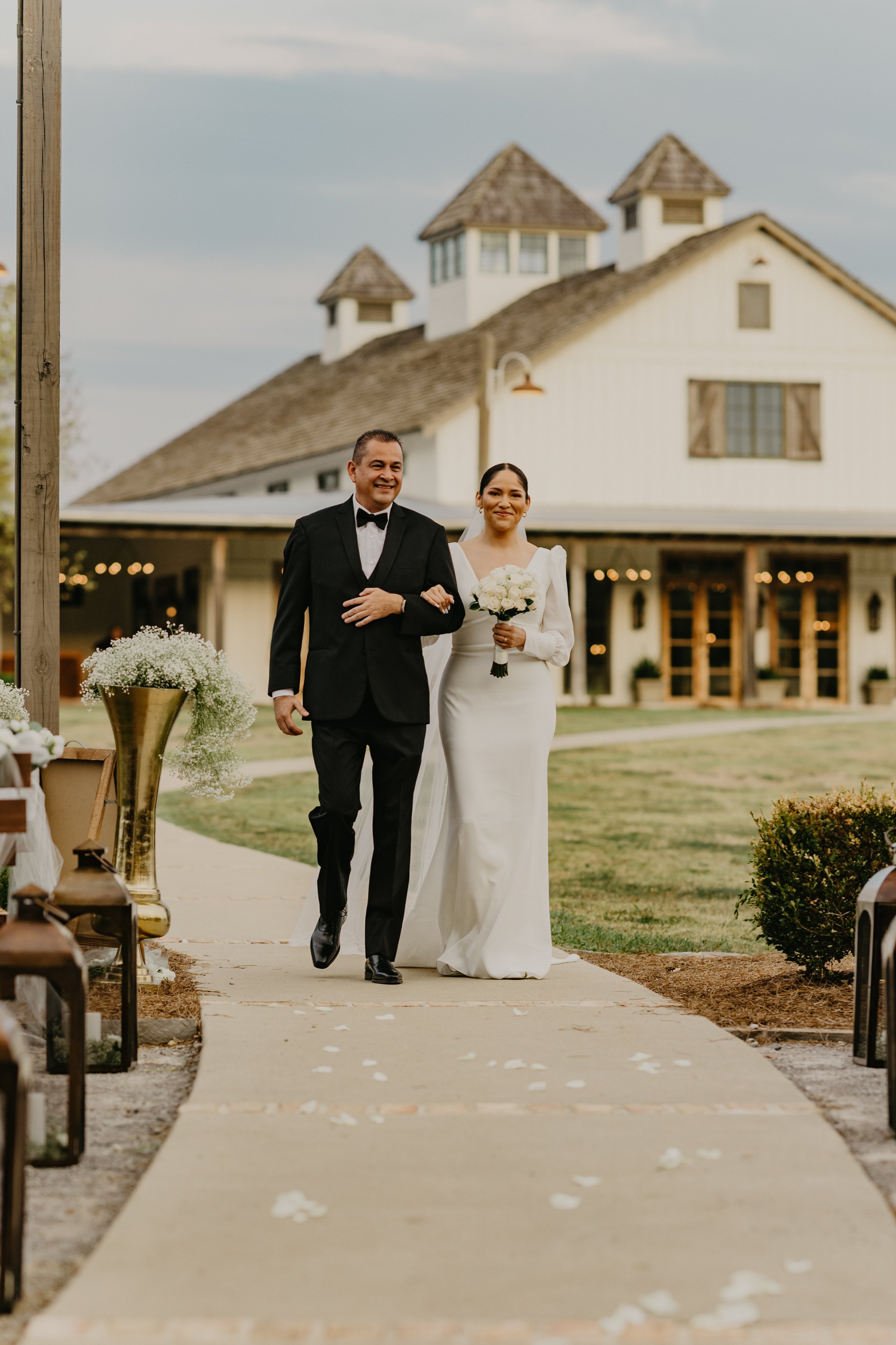 Bride walking down the aisle with father at outdoor wedding ceremony, with a rustic barn in the background.