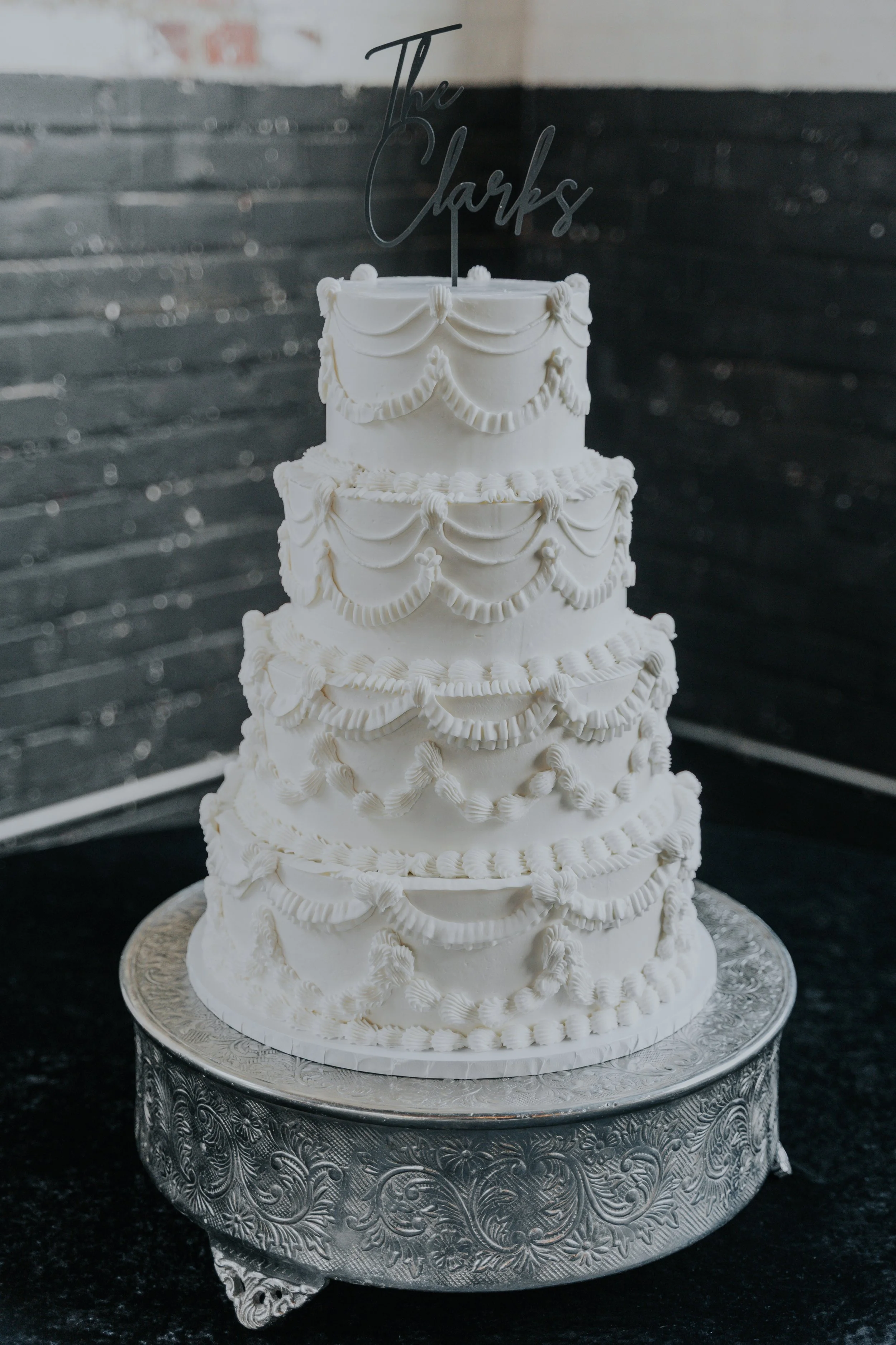 White four-tiered wedding cake with decorative piping, topped with a black 'The Parks' cake topper, on an ornate silver cake stand.