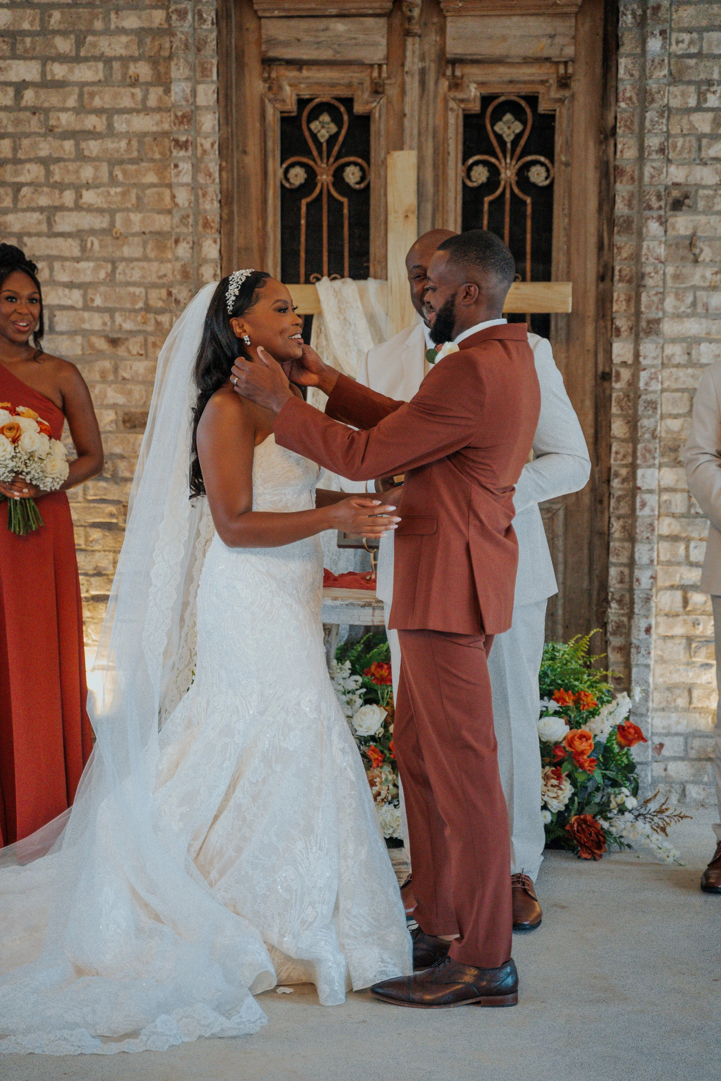 Bride and groom holding hands during a wedding ceremony, exchanging vows, with an officiant smiling behind them. Bridesmaid holds flowers on the left, and a decorated cross with flowers is in the background.