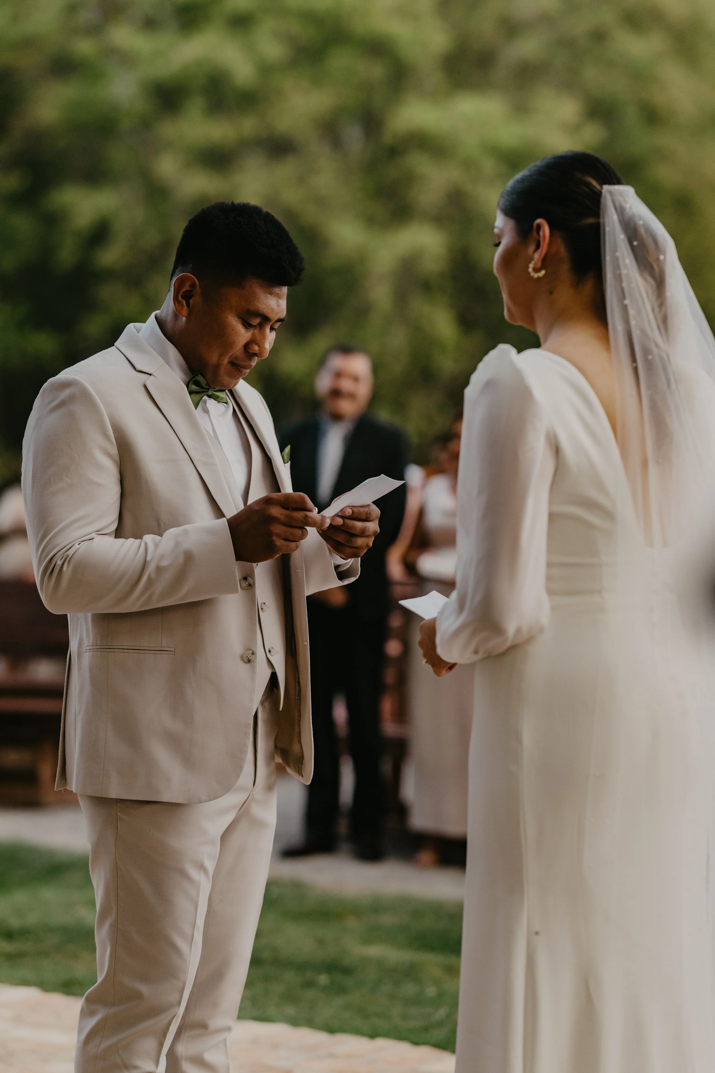 Bride and groom exchanging vows outside during a wedding ceremony, with a man reading from a note and guests in the background.