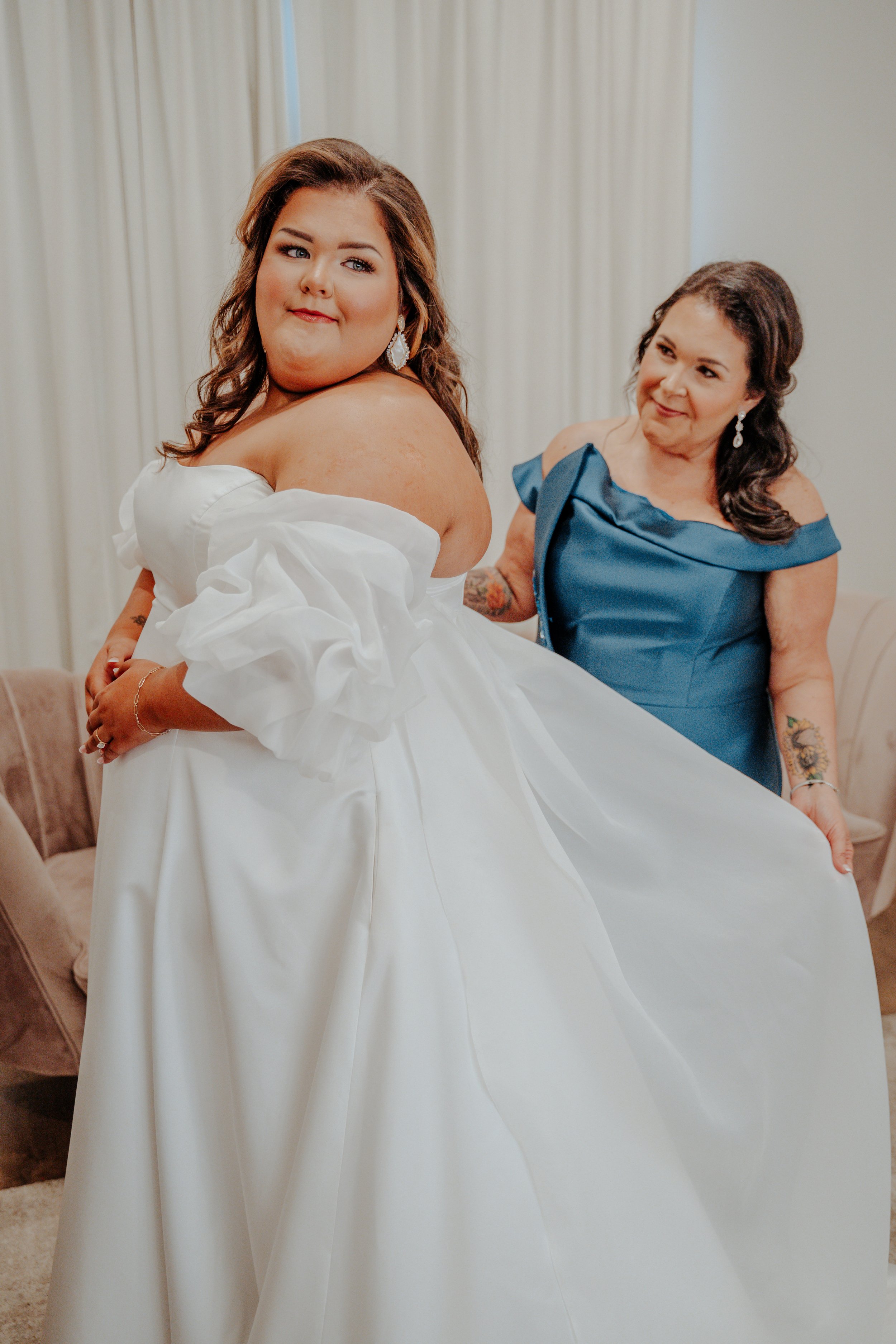 A bride in a white wedding gown with puffed sleeves is standing in front of a woman in a blue dress, both smiling softly in a well-lit room.