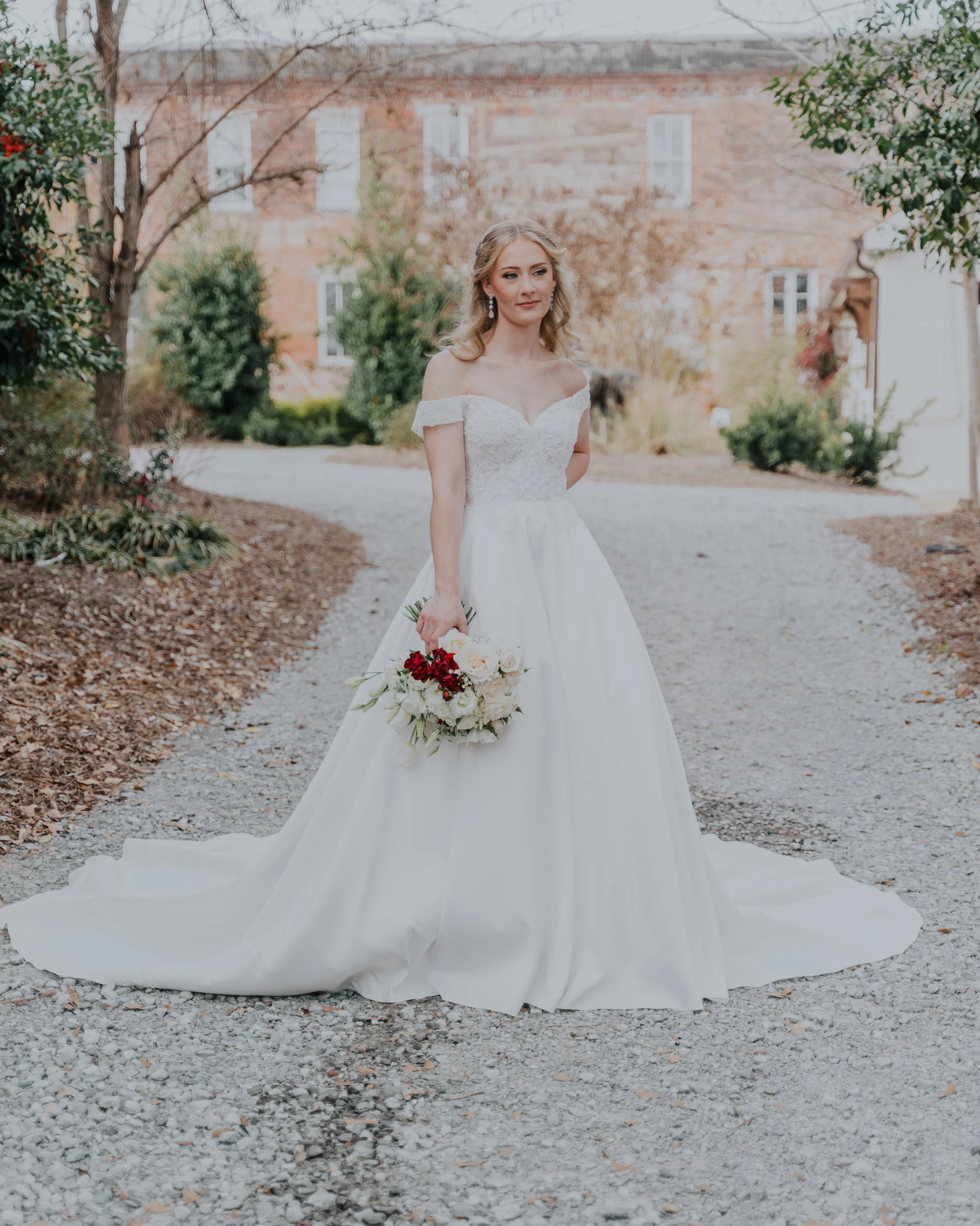 A bride in a white wedding gown holding a bouquet of white and red flowers outdoors on a gravel path, with greenery and a brick building in the background.