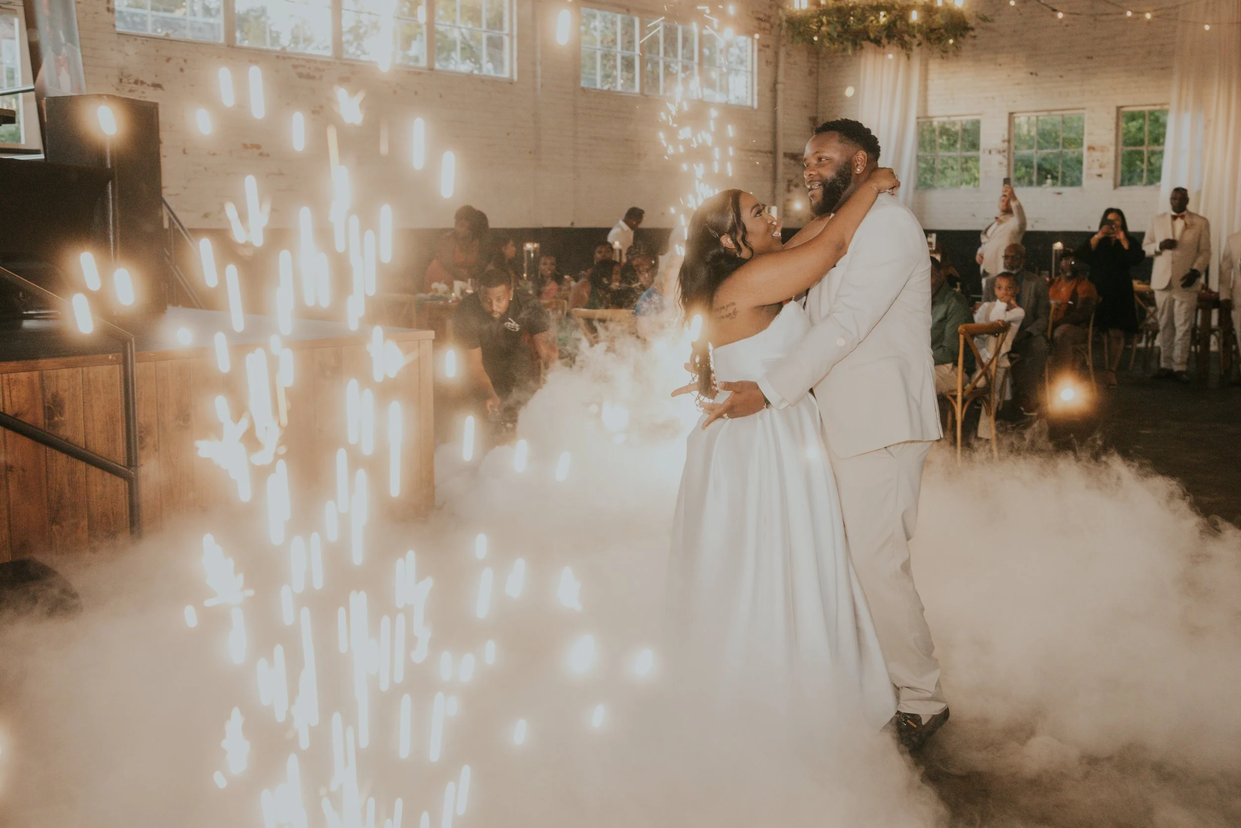 A newlywed couple sharing their first dance at a wedding reception, surrounded by guests, with special effects like fog and fireworks in a rustic indoor venue.