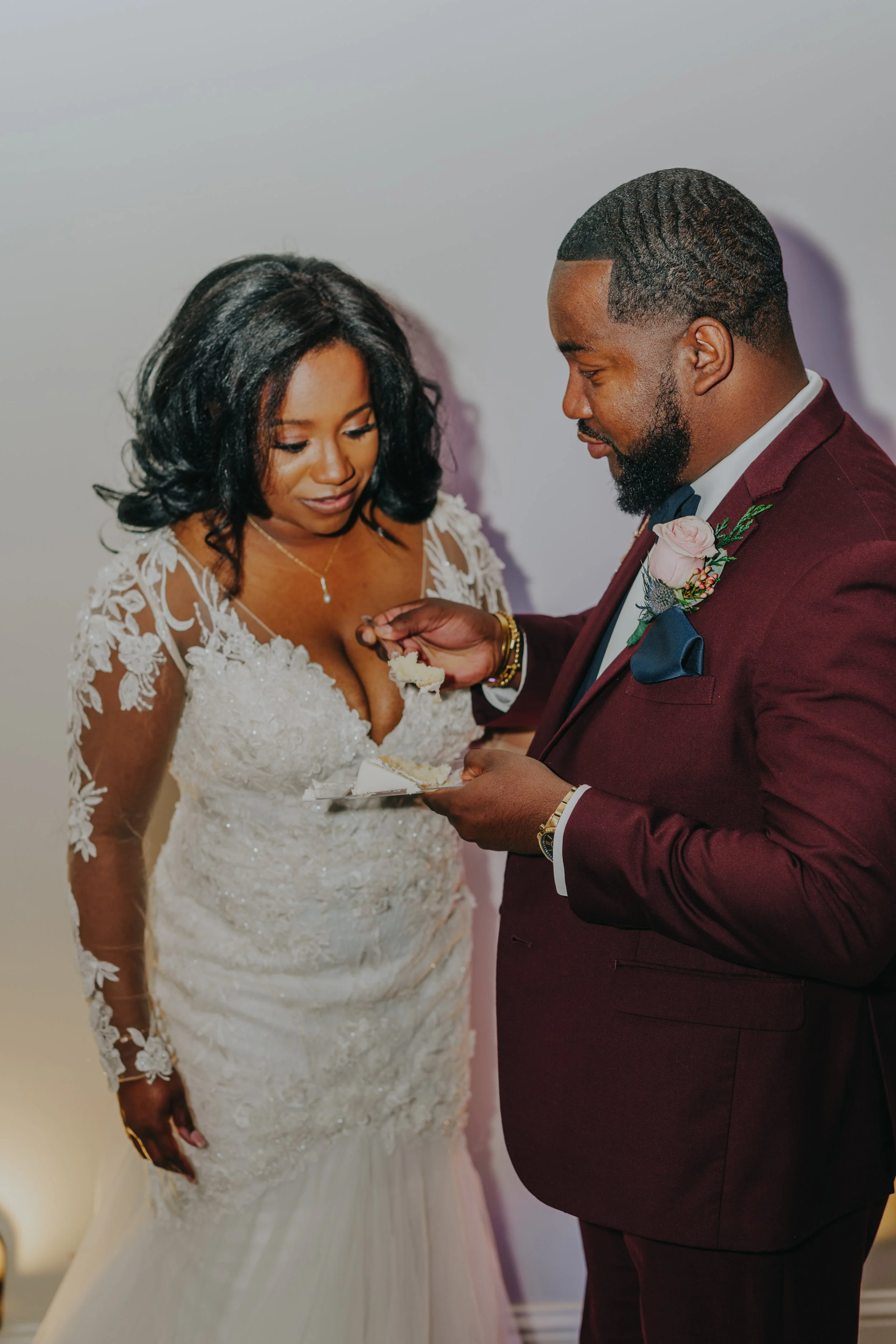 A man in a burgundy suit is feeding a slice of wedding cake to a woman in a white lace wedding dress during a wedding celebration.