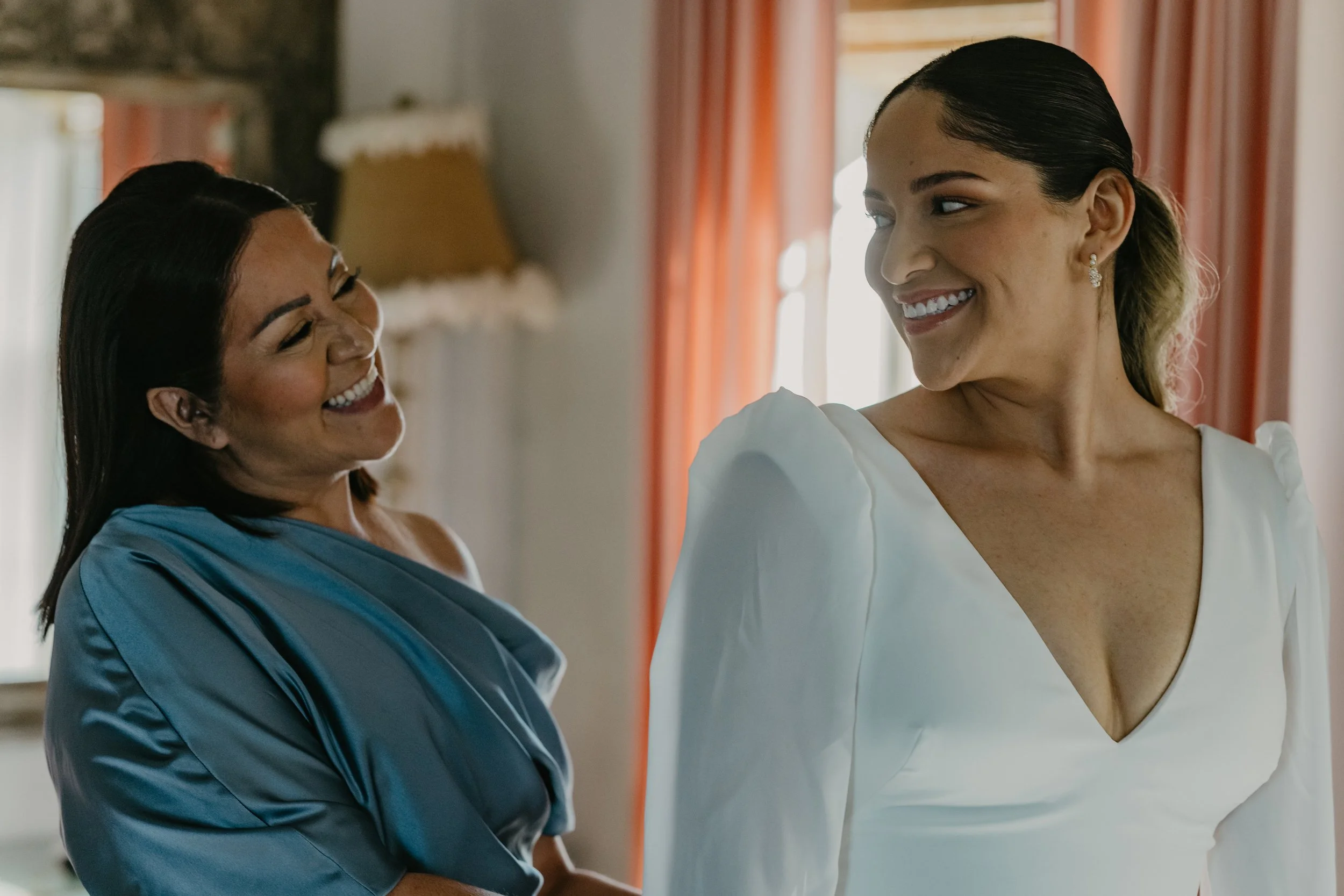Two women in a warmly lit room, smiling and looking at each other. One woman is wearing a blue dress and the other a white dress with puffed sleeves.