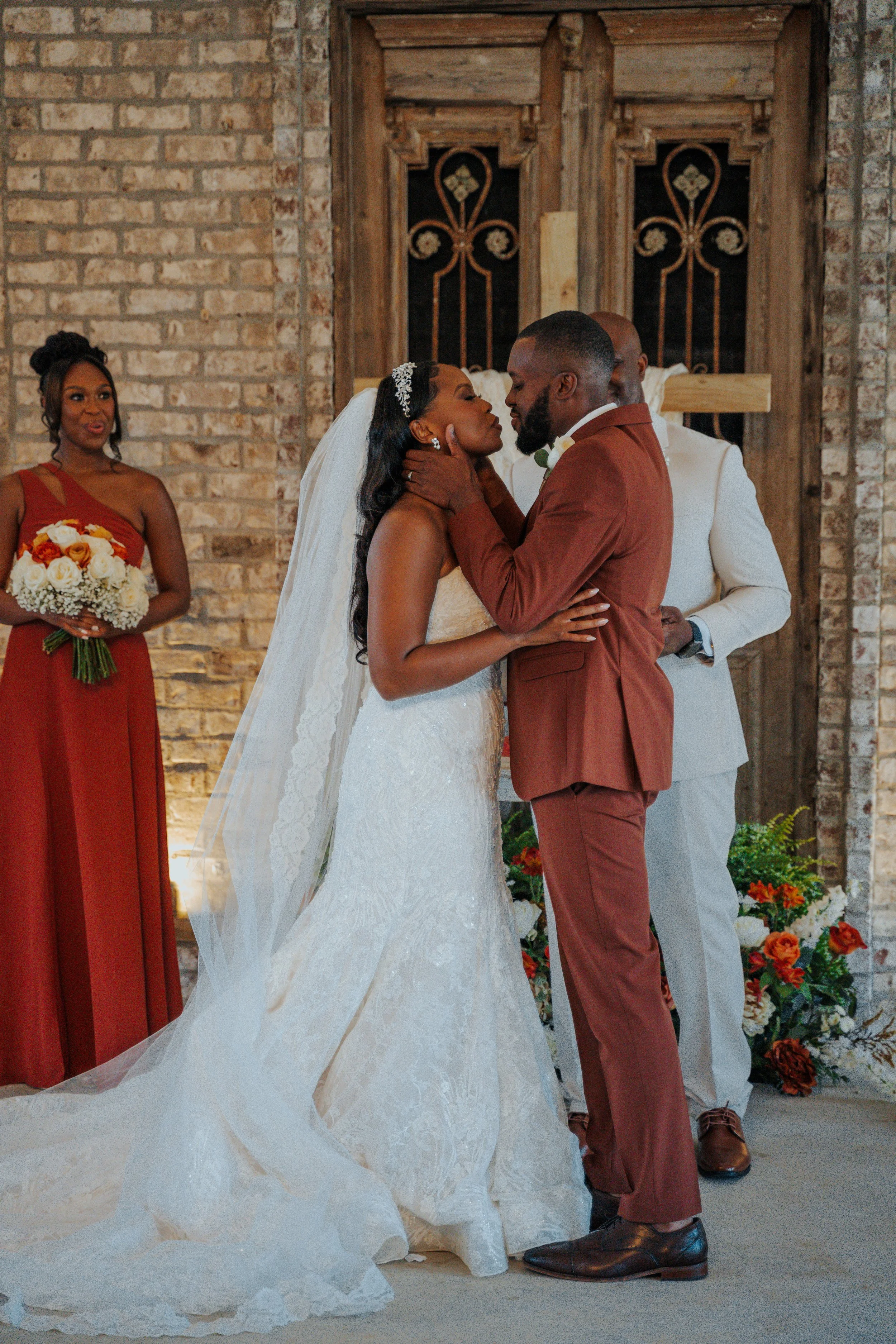 A bride and groom are about to kiss during their wedding ceremony, with a woman in a red dress holding a bouquet of flowers and a man in a white suit standing behind them, against a brick wall and wooden doors.