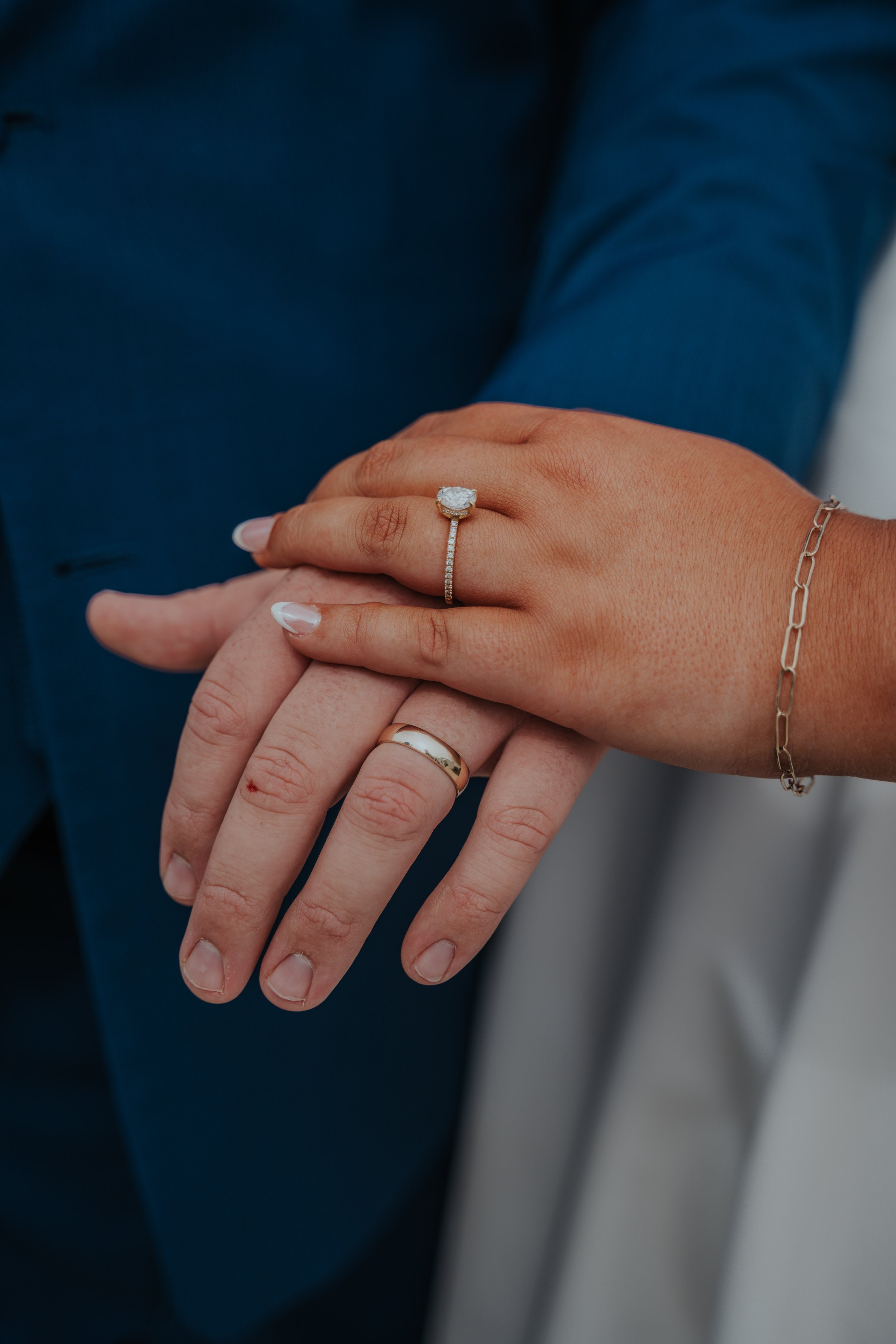 Close-up of a man and woman's hands with wedding rings, the woman wearing a diamond engagement ring and a silver bracelet, and the man wearing a gold wedding band, hands resting on each other.