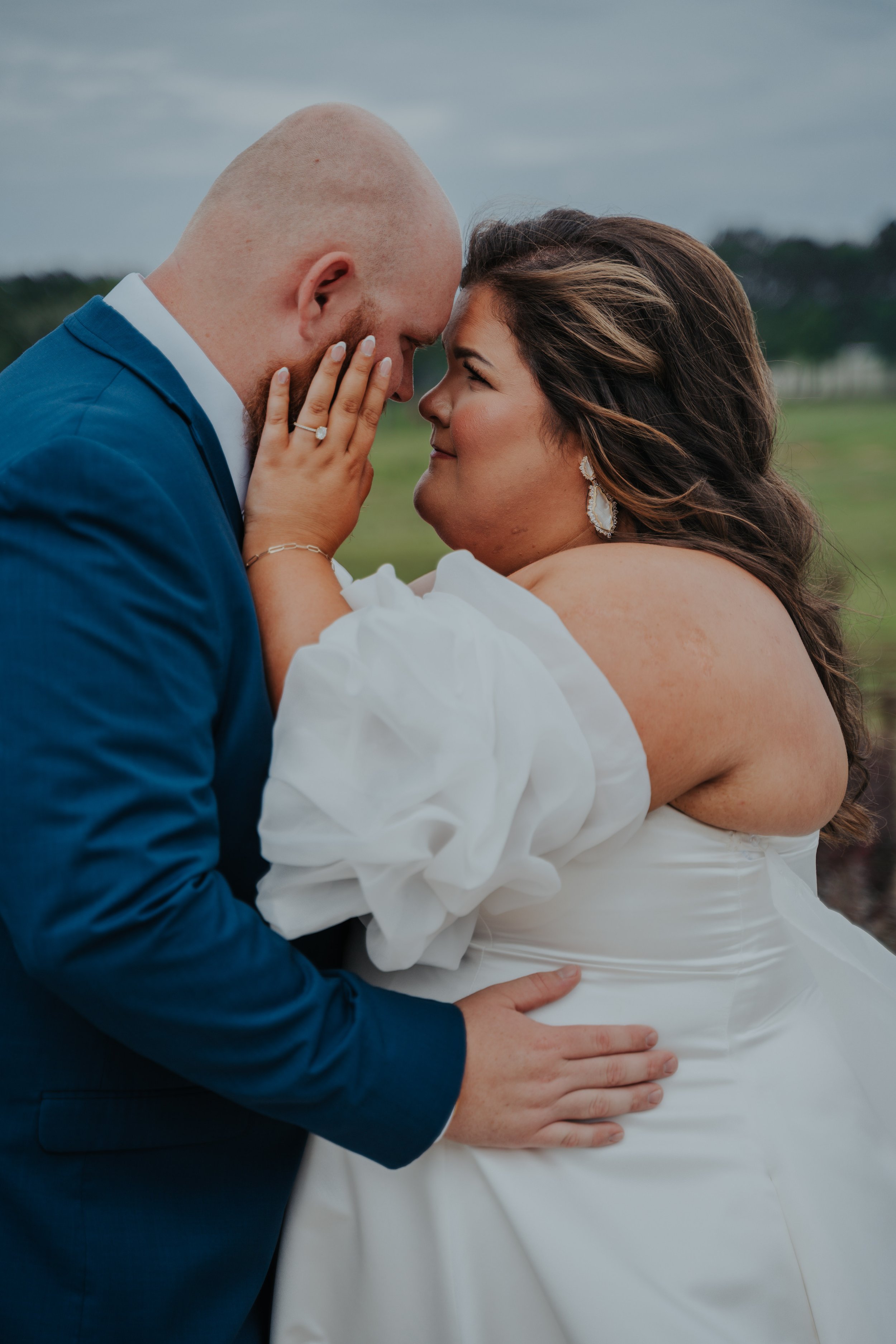 A couple sharing an intimate moment outdoors, with their foreheads touching and eyes closed. The woman has dark hair with highlights, wearing earrings and a white off-shoulder dress. The man is bald with a beard, dressed in a blue suit and white shir