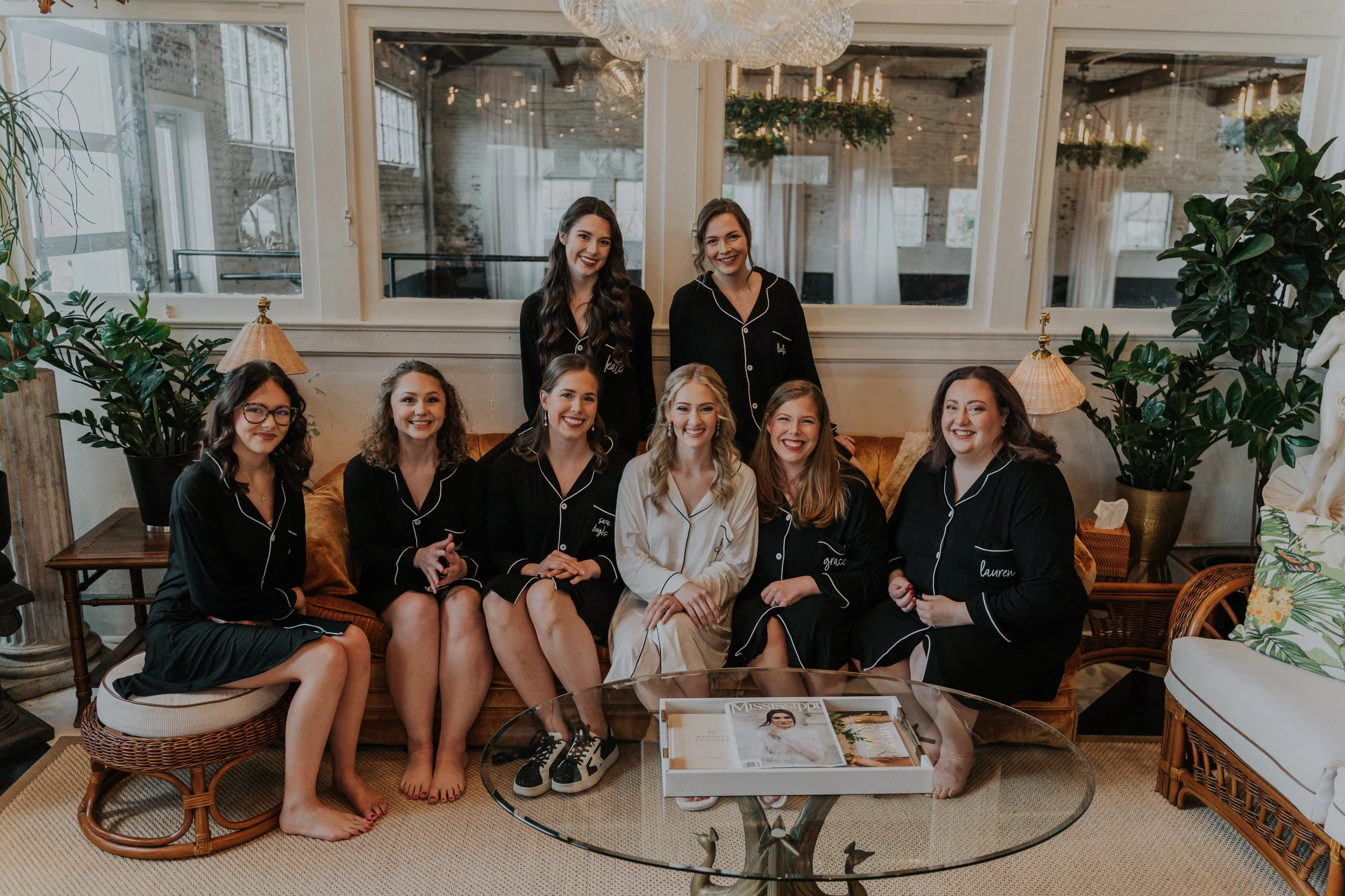 Group of nine women, some wearing black pajamas, sitting and standing on a vintage sofa and chairs in a bright living room, smiling at the camera, with greenery and large windows in the background.