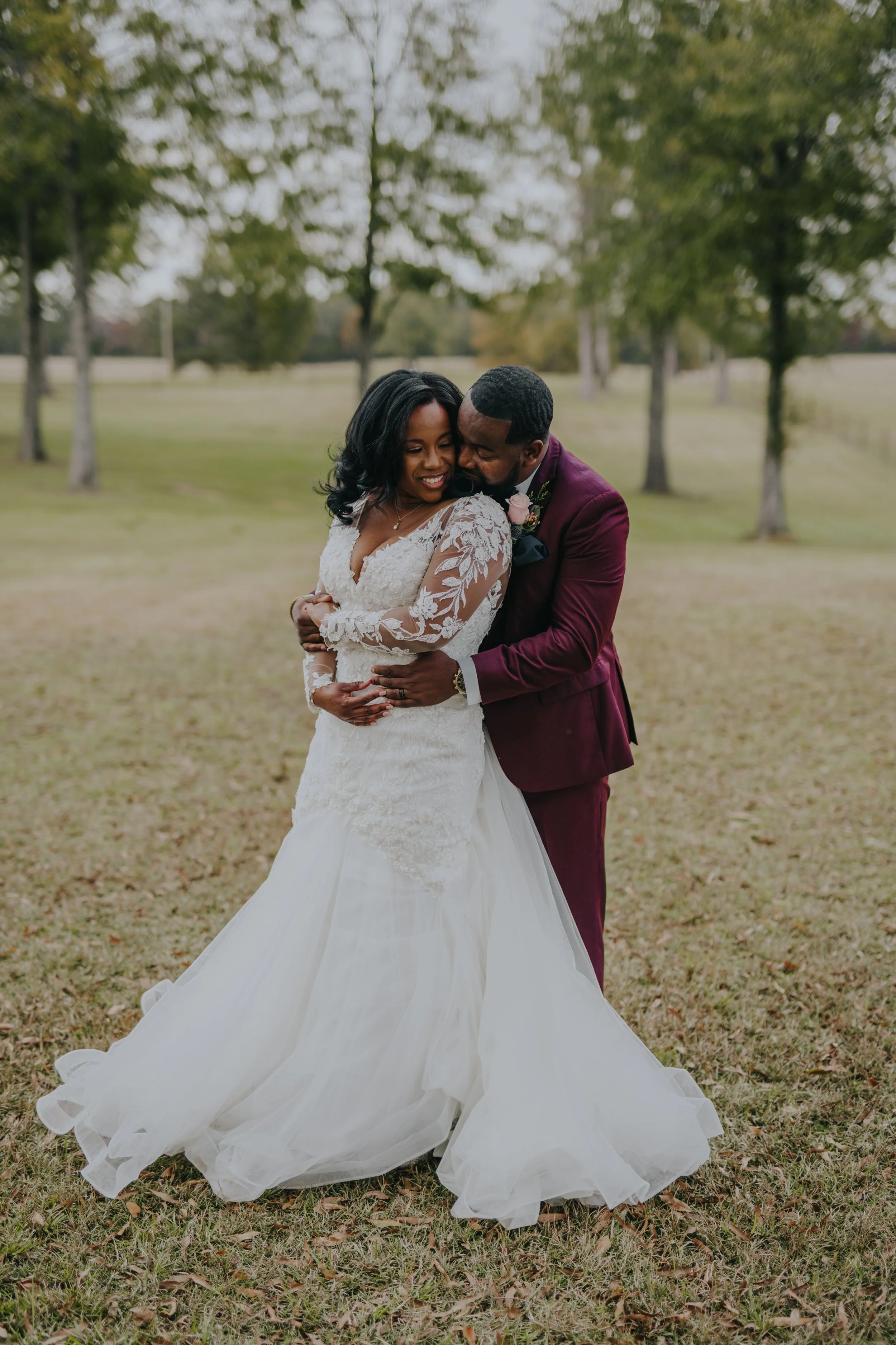 A couple in wedding attire embracing outdoors amidst trees, with the bride in a white lace gown and the groom in a burgundy suit.