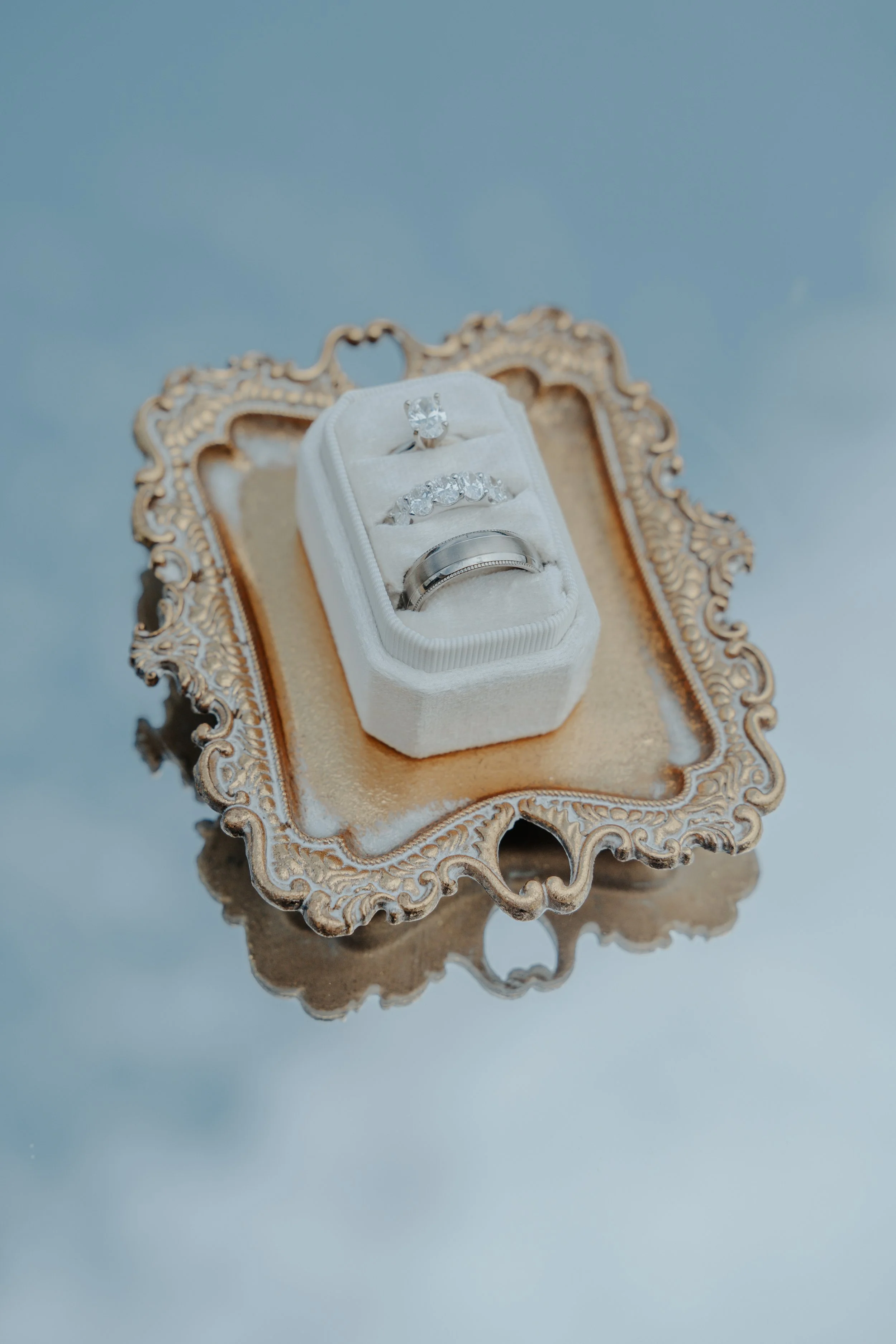 Three wedding rings, one solitaire and two bands, displayed in a white velvet ring box on an ornate silver tray.