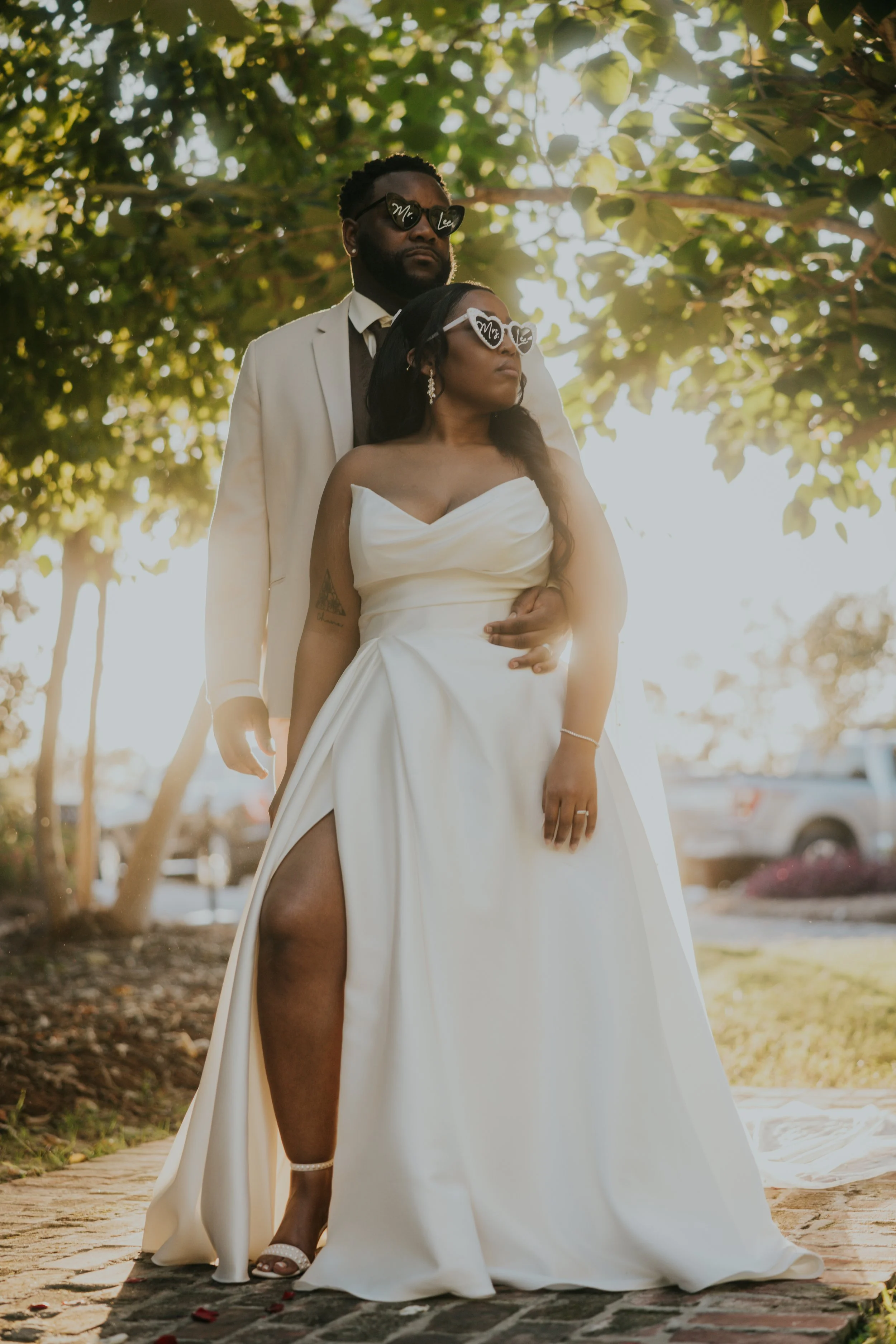 A couple in wedding attire standing outdoors under a tree, wearing sunglasses with personalized or playful designs.
