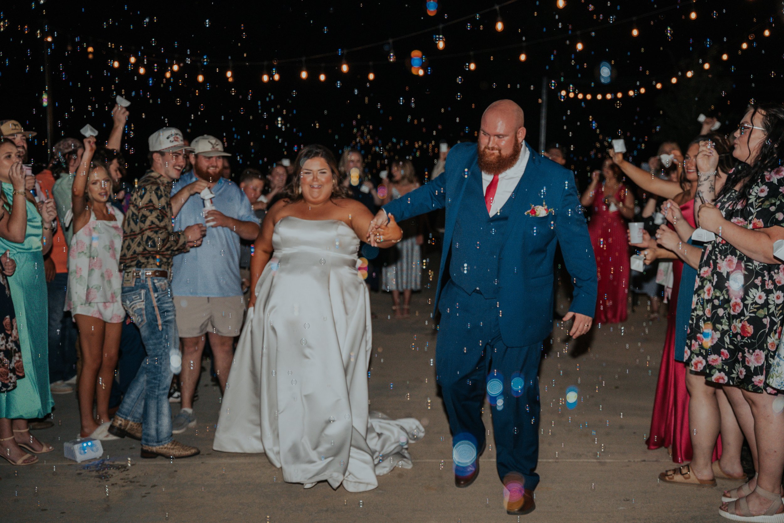 Bride and groom holding hands at night wedding celebration with guests, string lights, and confetti