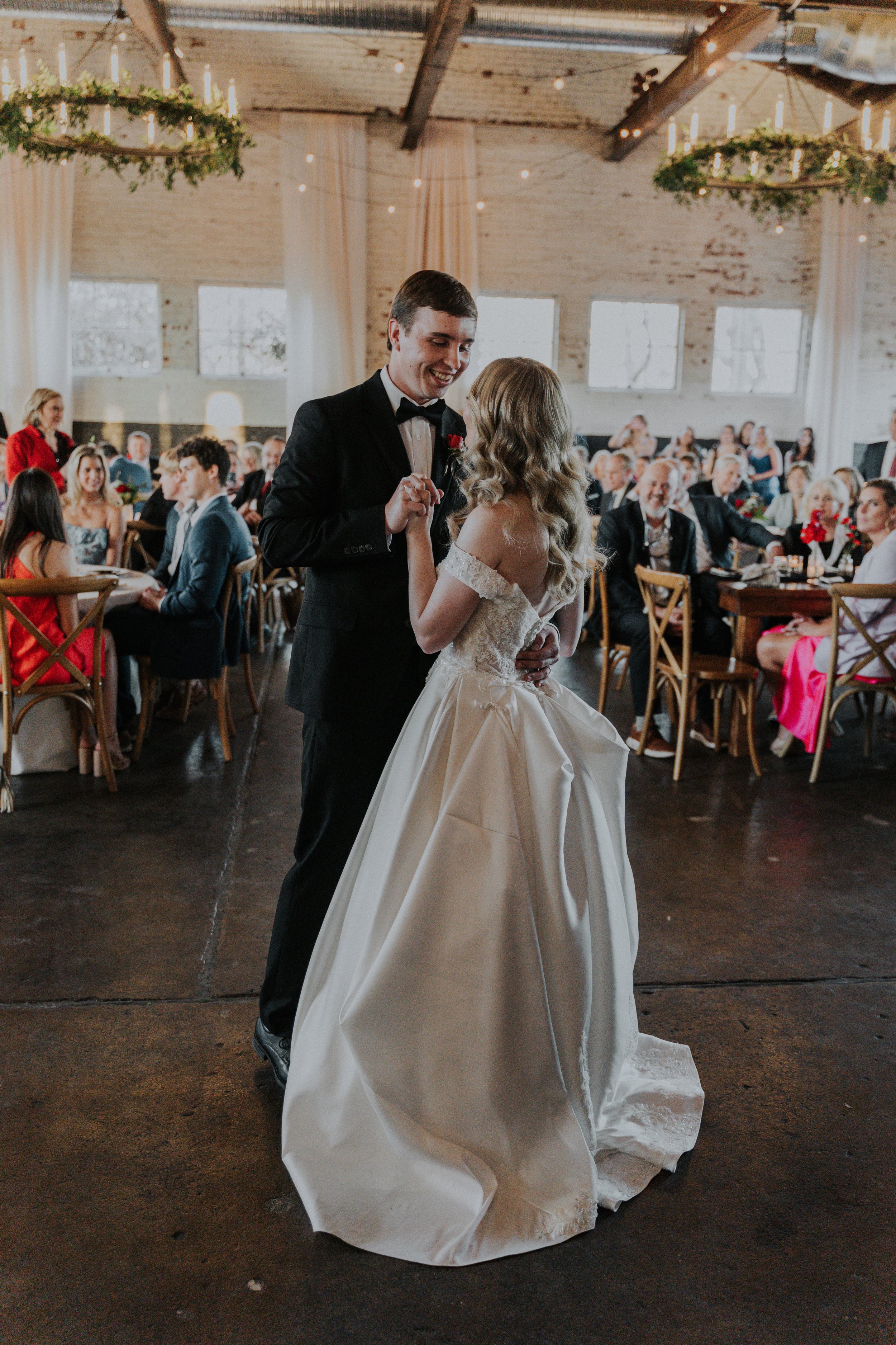 A newlywed couple shares their first dance at their wedding reception, surrounded by seated guests in a rustic indoor venue decorated with greenery and lights.