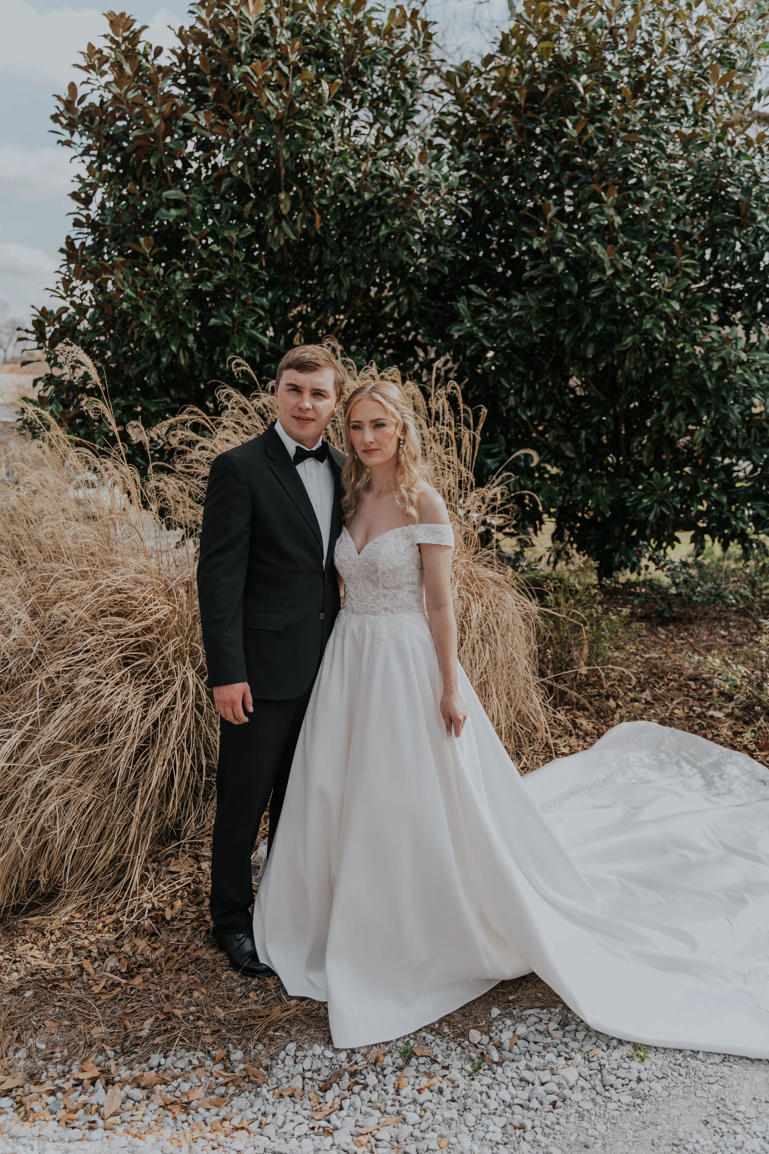 A bride and groom on their wedding day standing outdoors in front of a large green bush and tan ornamental grass. The bride wears an off-the-shoulder white wedding gown, and the groom wears a black tuxedo with a bow tie.