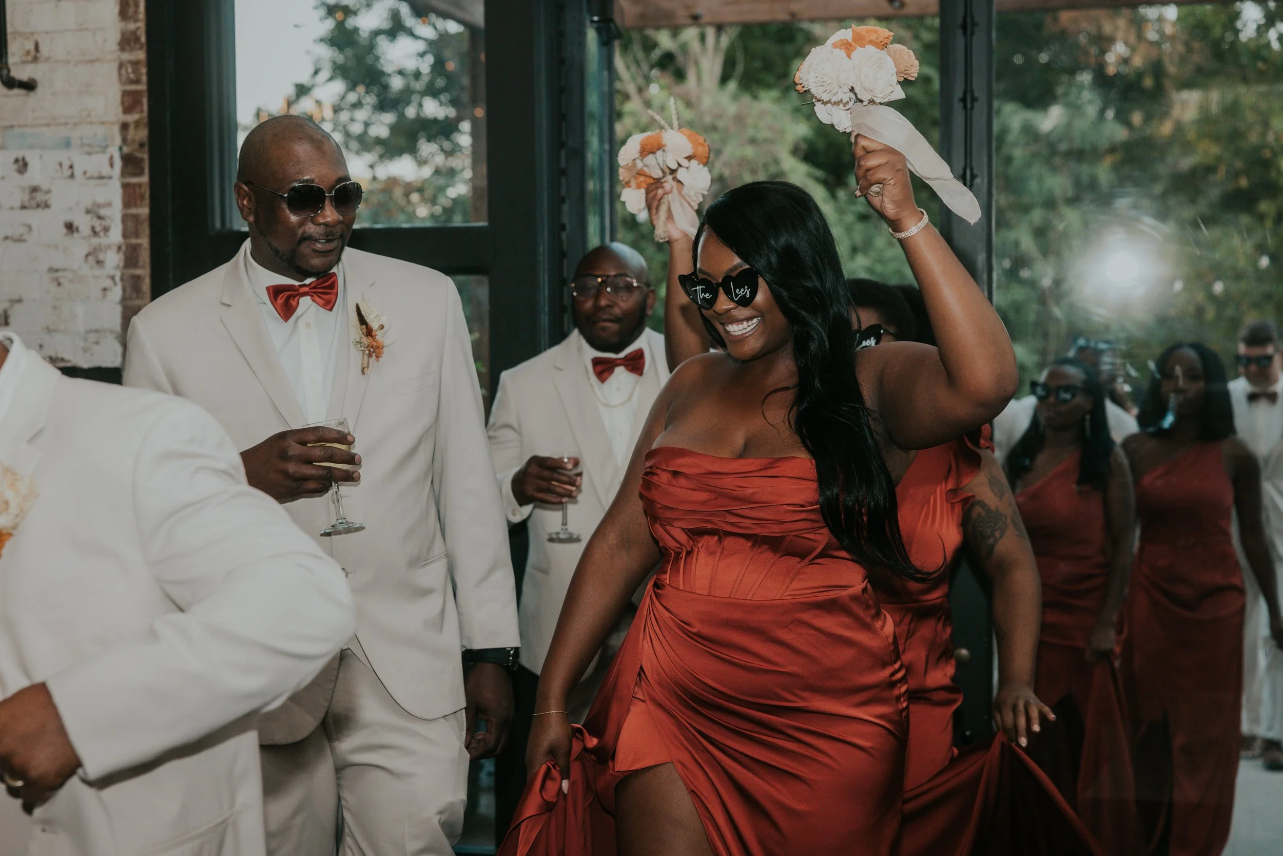 A group of people at a celebration, with a woman in a strapless red dress smiling and dancing, wearing sunglasses with the words "The Lees" on them, holding floral decorations above her head, surrounded by men and women in formal attire.